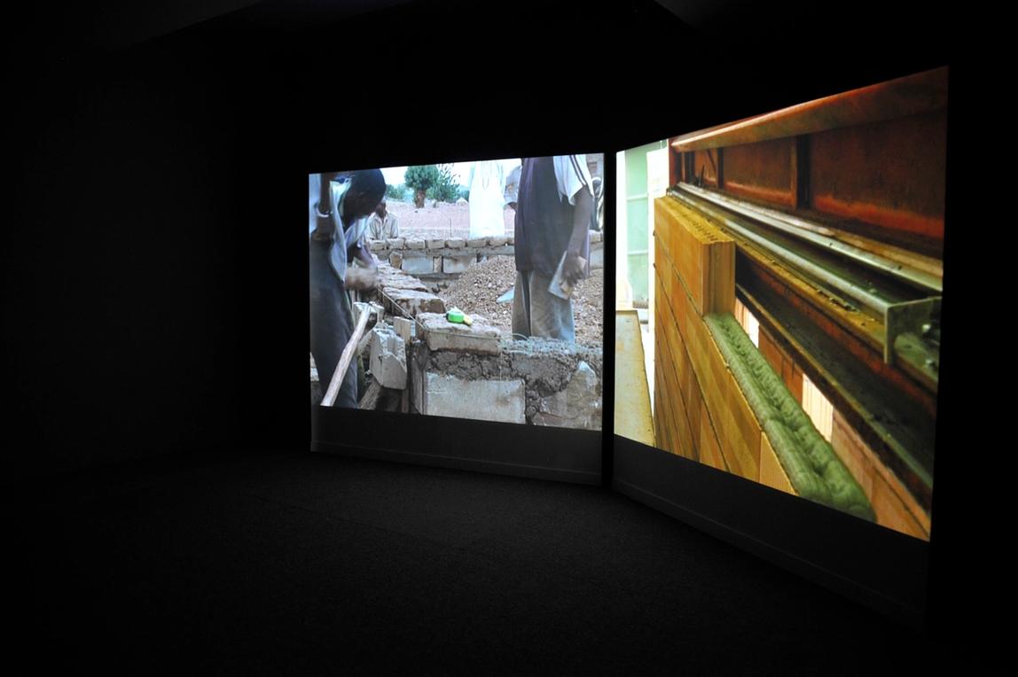 A two-channel video projection installed in a dark projection room. The left screen shows a group of people creating building blocks/plinths. The right screen, portrays a more automated manufacturing process of producing red bricks.