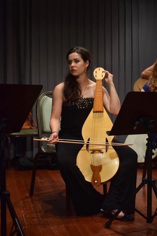 A young woman in black evening wear seating on a chair on stage and playing the fiddle.