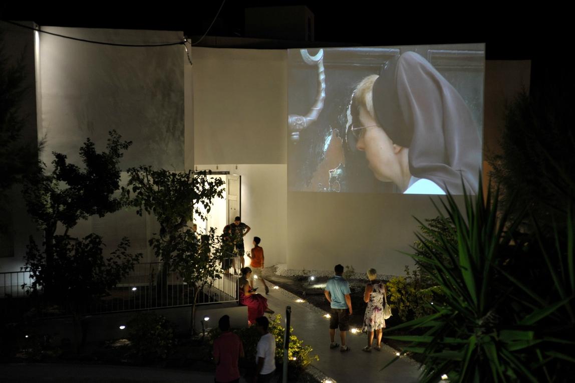 A wall projection on the Art Space Pythagorion's facade at night showing a woman, possibly a nan looking through a hole on a door. The building is surrounded by vegetation. A group of people is standing in front of the building looking at the projection.