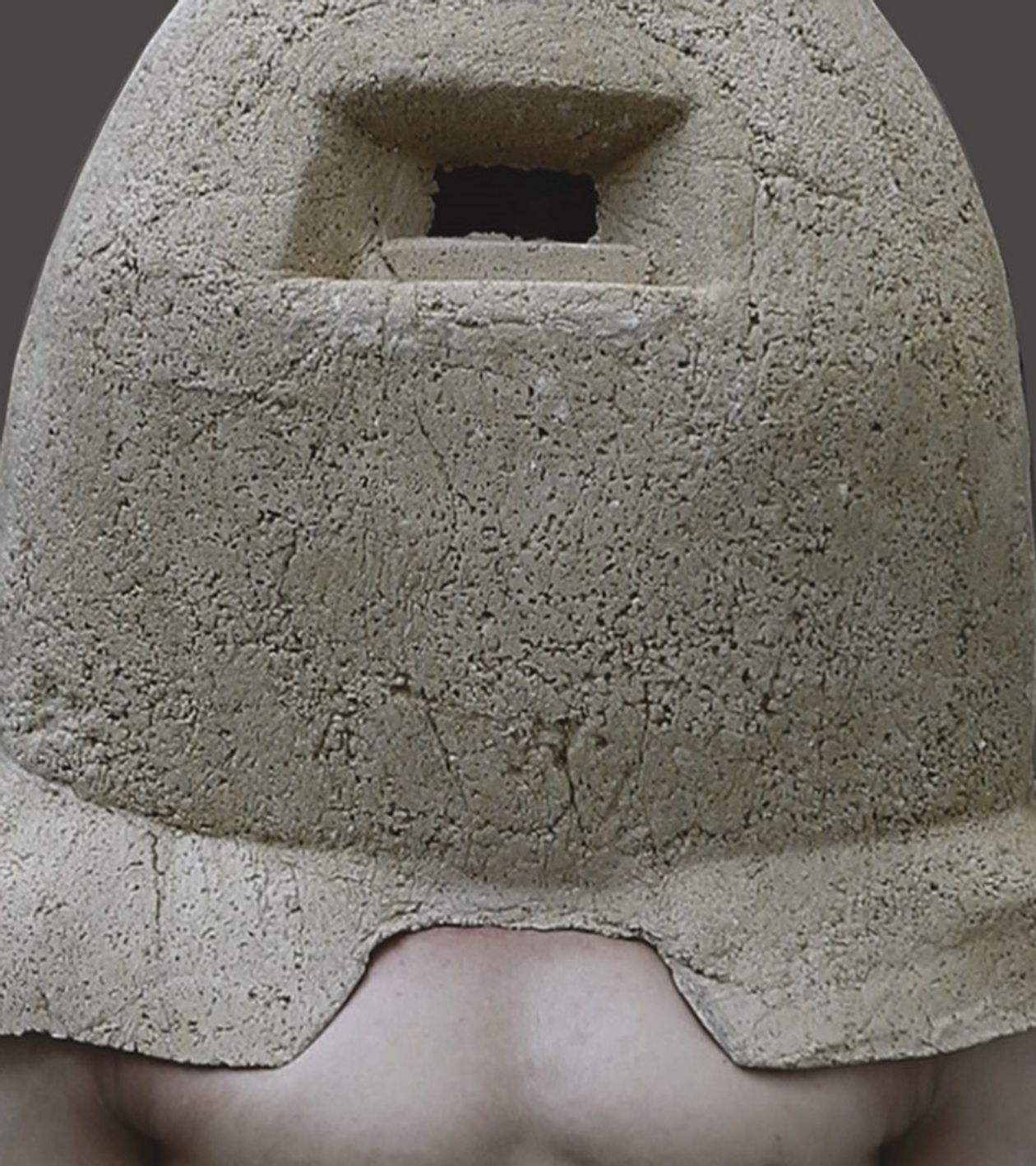 Portrait of a naked man. His torso is visible but his head and shoulder are hidden under a helmet-like structure of limestone. 