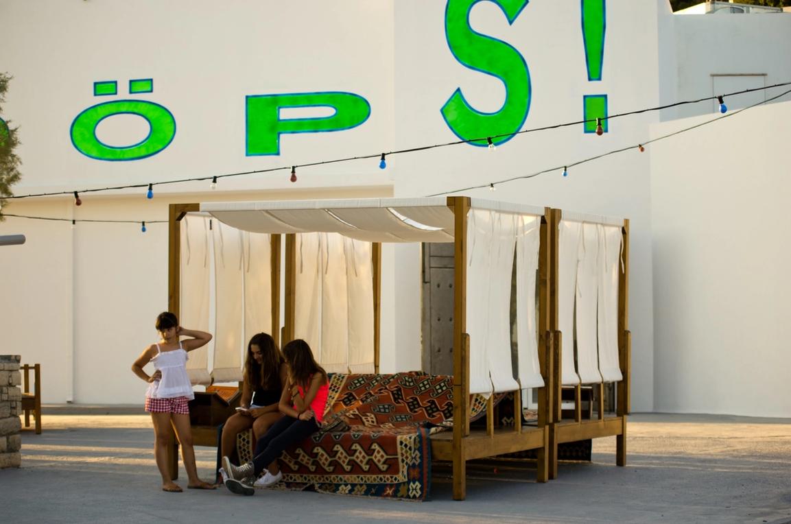 Children are seating and interacting on large wooden kilim beds place in Art Space Pythagorion's courtyard. In the background, the building's white walls feature the word "ööööööps!" in bright green letters.