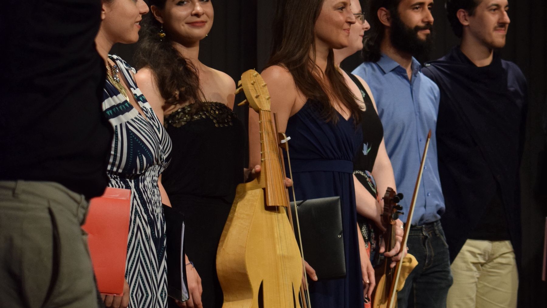 A group of people of different ages on stage arranged in two rows. In the front row 8 people are seating, each one playing a baroque instrument, i.e the flute, the lute, the recorder etc. Six people standing behind them are holding envelopes and they are singing.