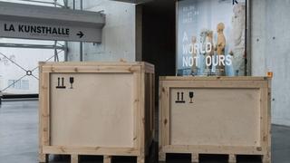 Two cargo wooden boxes inside a contemporary building. A banner of the exhibition "A World Not Ours" hangs on one side of the wall, in the background