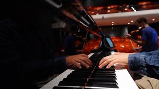 Εστίαση στα χέρια ενός πιανίστα και τα πλήκτρα ενός πιάνου σε μια άδεια αίθουσα. Στο φόντο ένας βιολιστής κάνει πρόβα με το βιολί