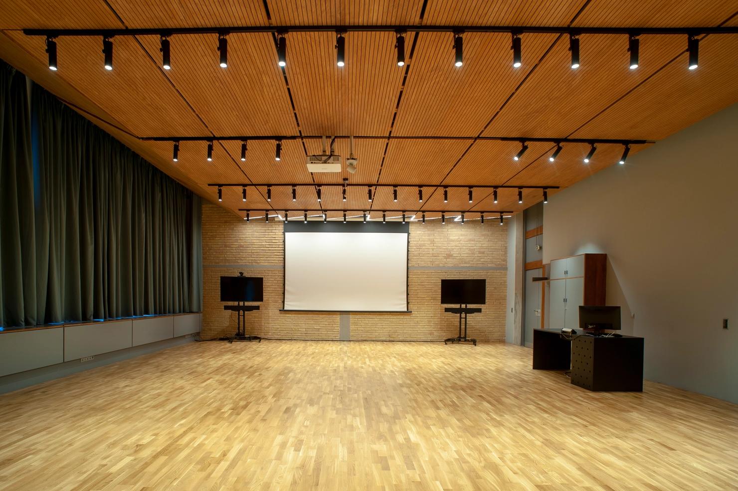 The distance learning studio is a room within the Athens Conservatoire building with wooden flooring and ceiling. On a brick wall there are two monitors between a blank projection screen. To the left, there is a desk with a computer next to a standing cabinet.
