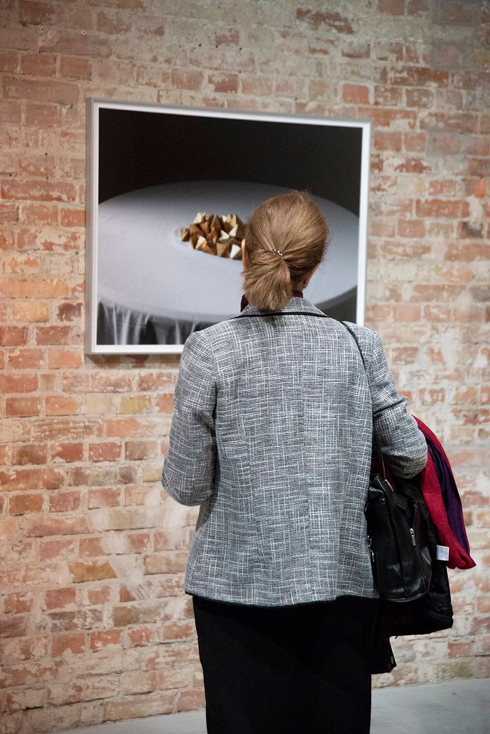 A visitor standing in front of a framed work hanging on a brick wall and looking. The work is a close-up of a pile of triangle-shaped individually wrapped chocolates on a surface. 