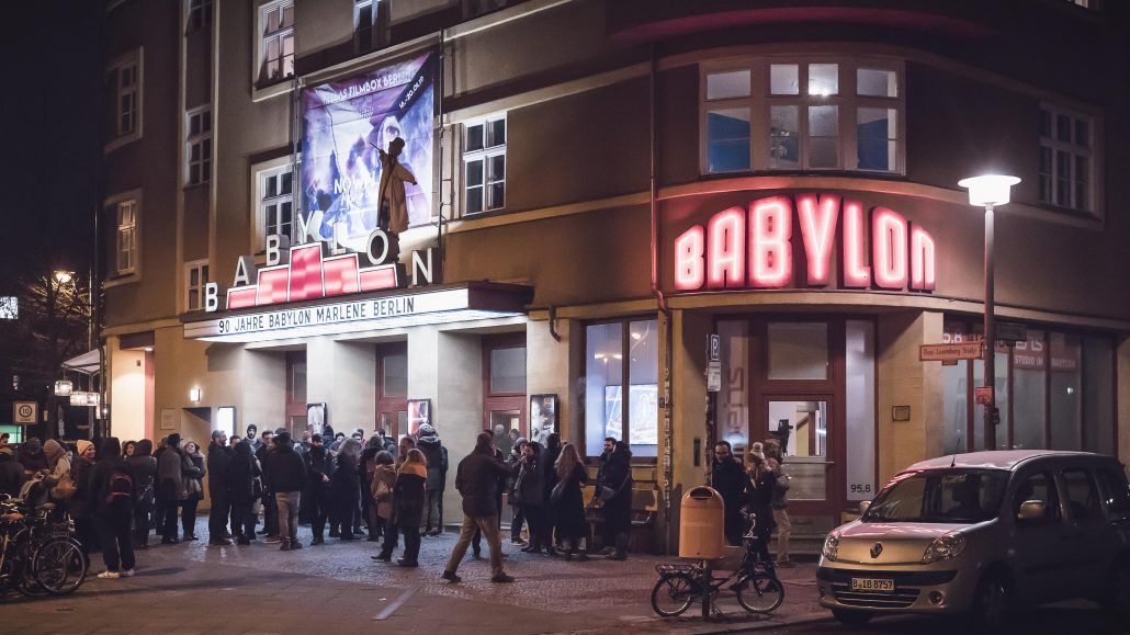 The facade of the arthouse cinema hall "Babylon" in Berlin. A group of people is standing in front and a large red neon sign features the word 'BABYLON"