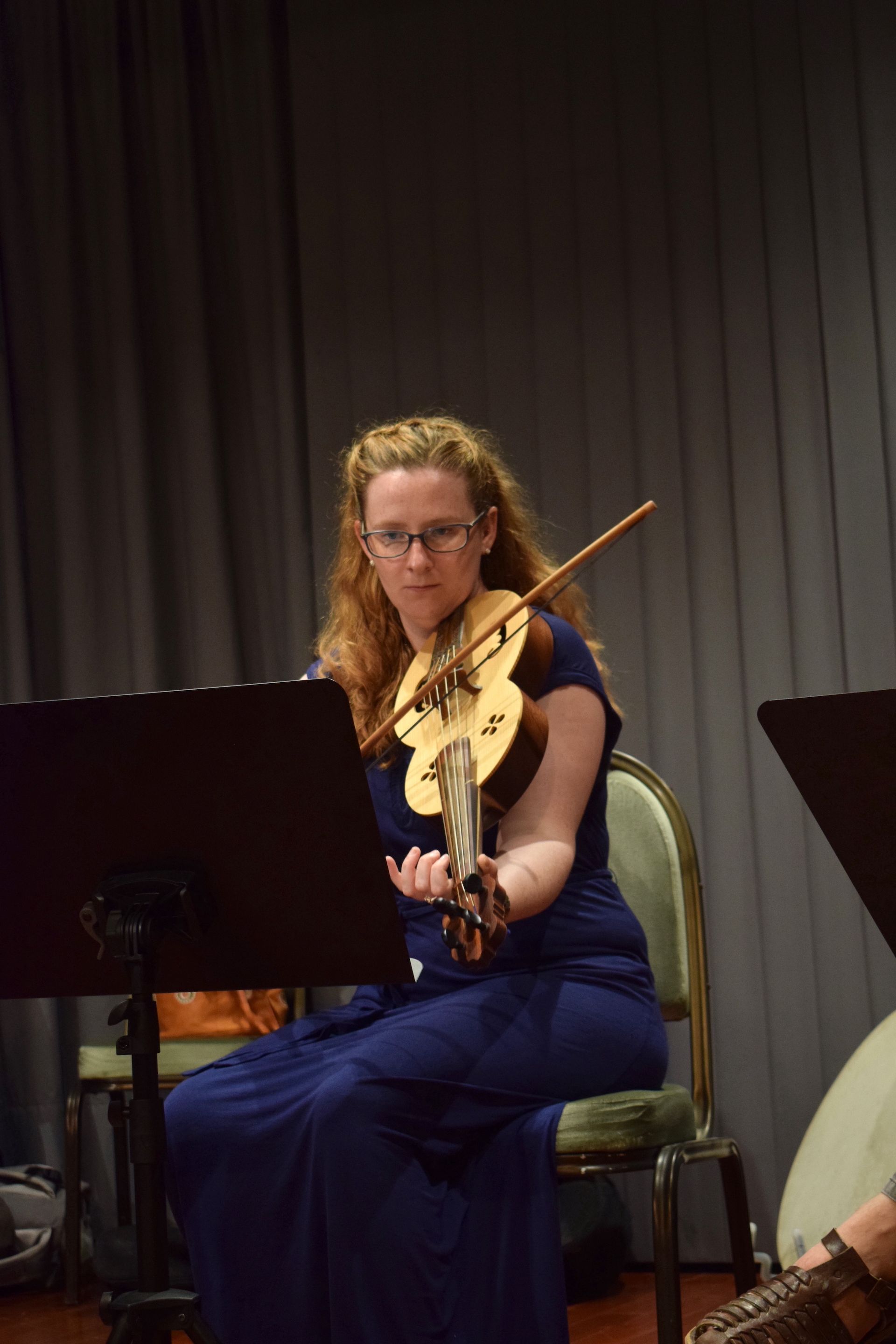 A young woman in blue evening wear seating on a chair on stage and playing the fiddle.