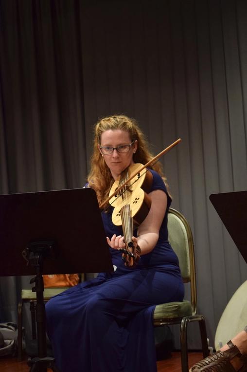 A young woman in blue evening wear seating on a chair on stage and playing the fiddle.