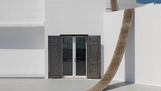 Ein langer, schmaler orientalischer Teppich hängt über der Fassade des Art Space Pythagorion. Er hängt von der Spitze des Gebäudes über die strahlend weißen Wände und endet im Innenhof des Gebäudes. Die doppelten Eisentüren des Art Space Pythagorion sind offen.