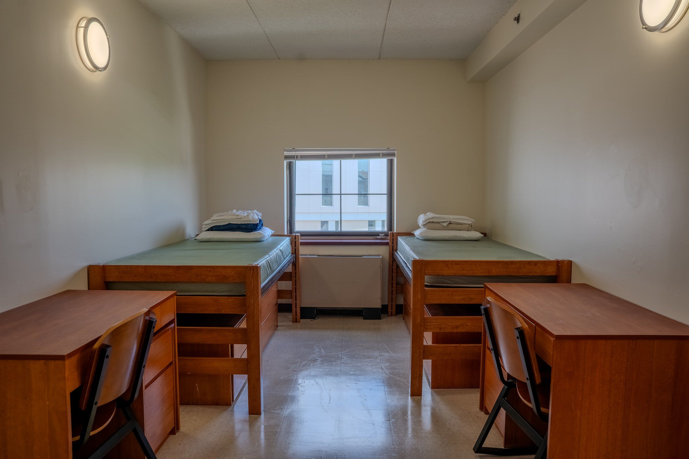 Two single beds with wooden desks and chairs under a window in a St. John's residence room