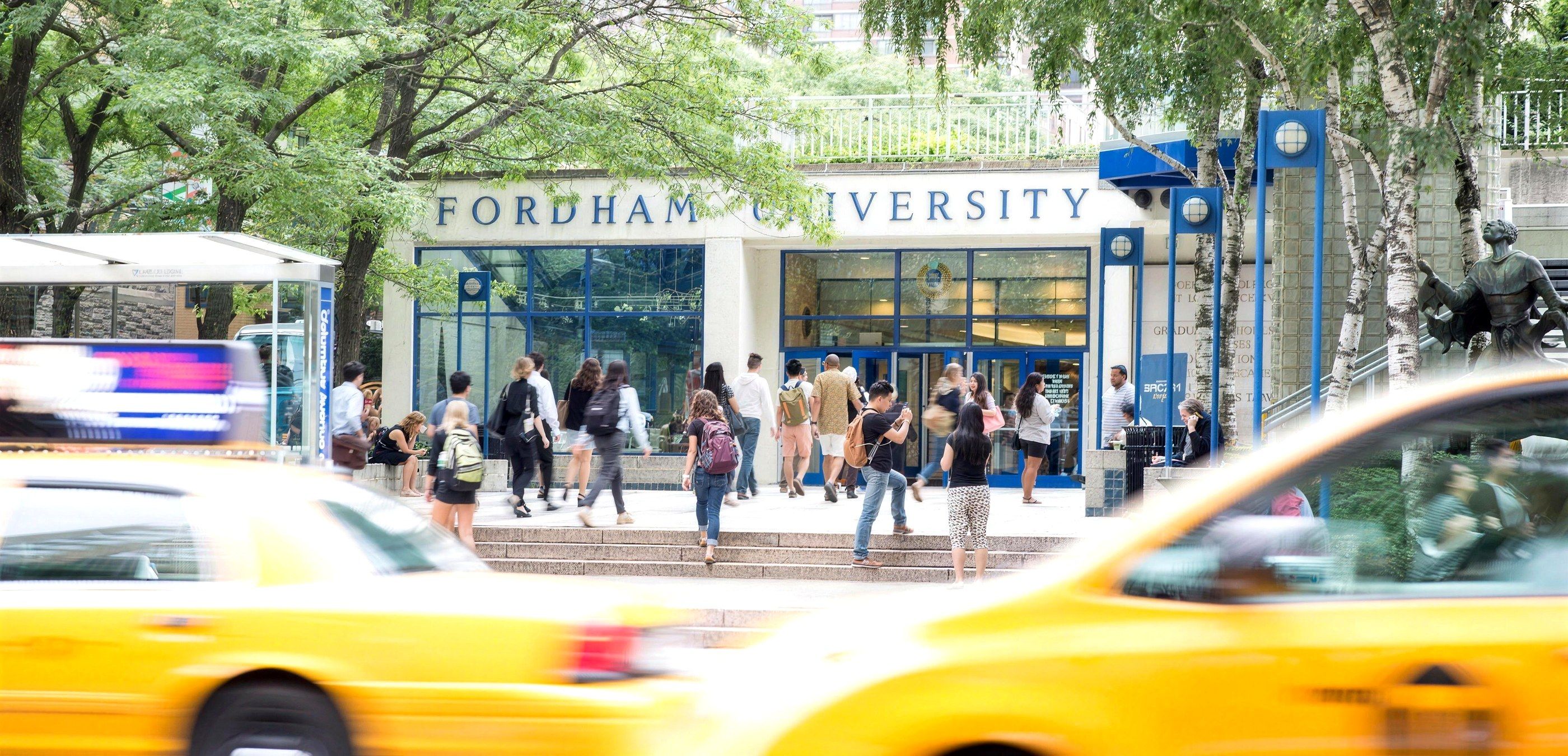Students walking into the Fordham University entrance at Lincoln Center, with yellow taxis passing