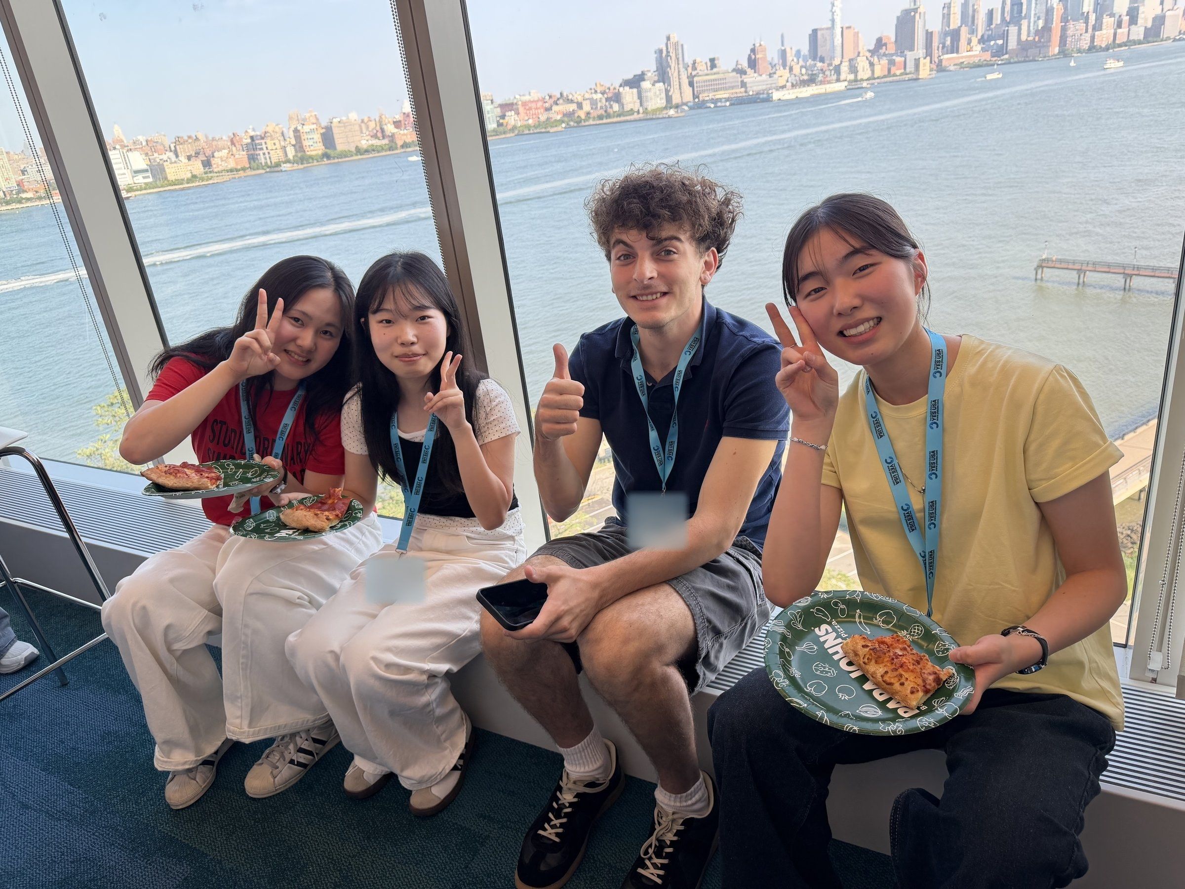 Smiling students with pizza by a window overlooking the Hudson and Manhattan