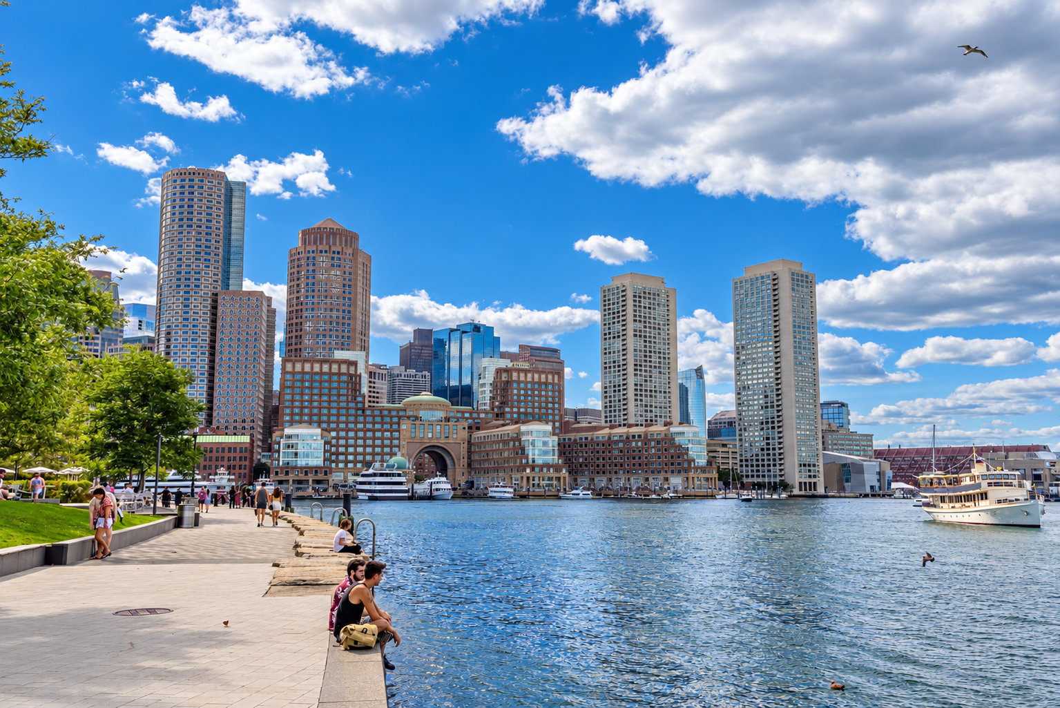 Boston waterfront skyline with blue skies, boats and a tree-lined promenade.