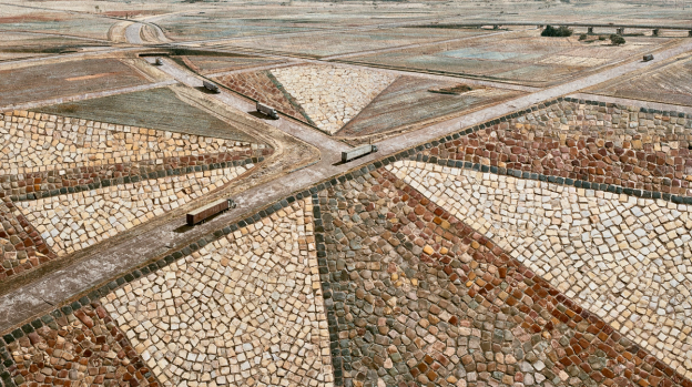 Aerial view of freight trucks travelling along intersecting dirt roads that cut through a vast yard of stone blocks laid out in triangular segments