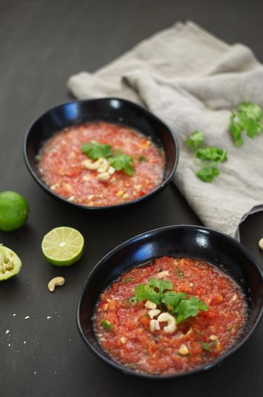 90 degree angle shot of two bowls filled with watermelon gazpacho, garnished with roasted cashews and coriander on a table with a linen cloth and lime halves