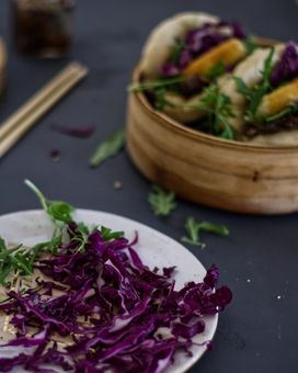 Plate with fresh cabbage and rocket, next to a steamer with bao buns (gua bao) with rocket, cabbage, and fried goats cheese