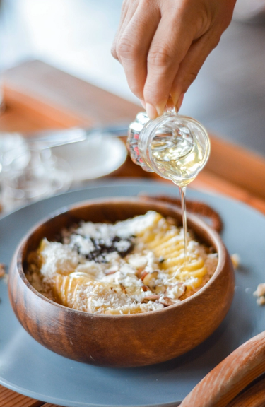 Porridge topped with fruit, nuts and coconut shavings in a wooden bowl with a hand drizzling honey from a little glass