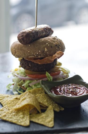 Head shot of a towering burger with Beyond Meat (Vegan) Sausage, Patty, vegan bacon, salad and a side of nachos with tomato sauce