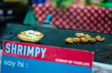 Grill with a burger bun besides a handful prawns and the logo of food stall, Shrimpy in London