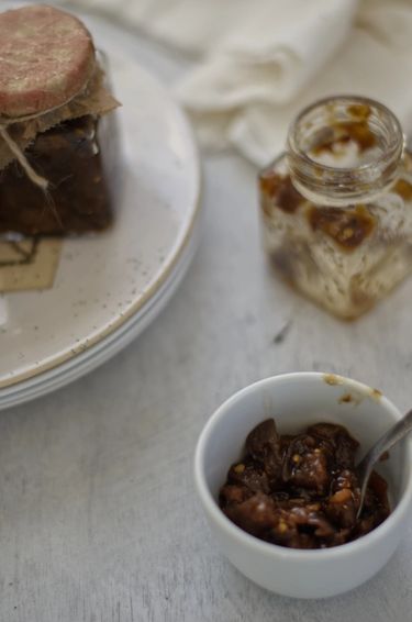 A close-up of a bowl of chutney surrounded by an empty glass jar and a full jar on a table with a white cloth