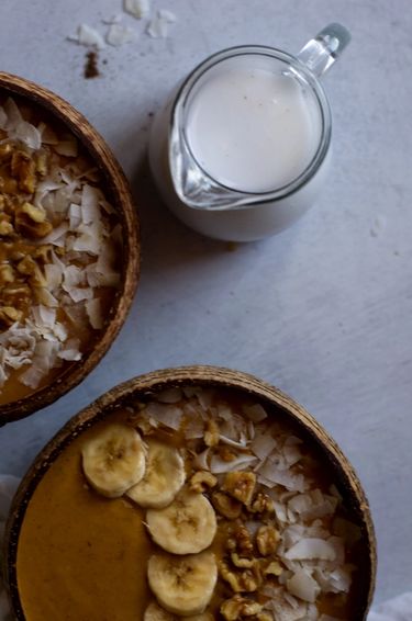 Top view of  two smoothie bowls topped with coconut shavings, walnuts and banana next to a jug of coconut milk 