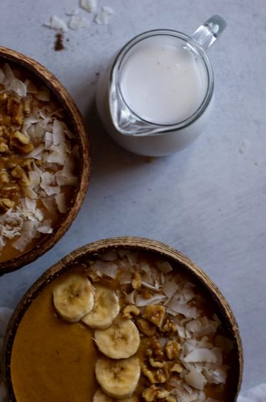 Top view of two smoothie bowls topped with coconut shavings, walnuts and banana next to a jug of coconut milk