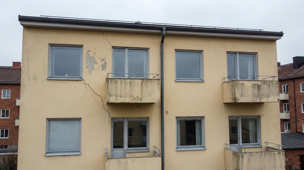 Drönarbild rakt mot en gulputsad fasad i höjd med övervåningen: en spricka i putsen som löper snett nedåt, färg som flagnat under takfoten, betongbalkonger med smutsränder och ett stuprör