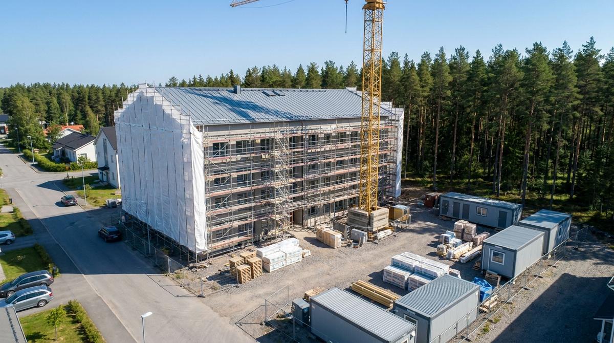 Flygbild av ett flerbostadshus under uppförande med tak och fönster på plats, inklätt i byggställning med vit väderskyddsduk, en tornkran, bodar och pallar med material på grusplanen
