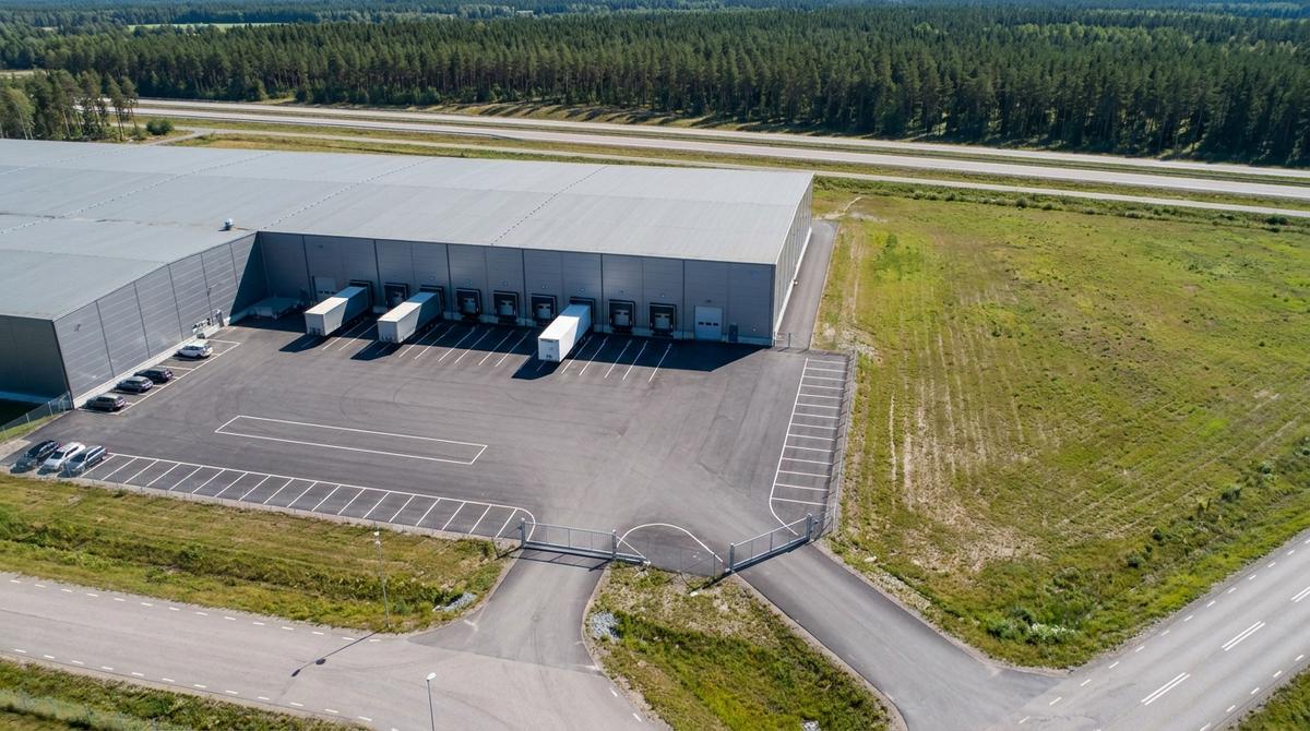 Flygbild av en lagerfastighet med lastkajer och lastbilssläp, asfalterad plan med parkering och infart med grind, en obebyggd tomt bredvid och motorväg och skog i bakgrunden
