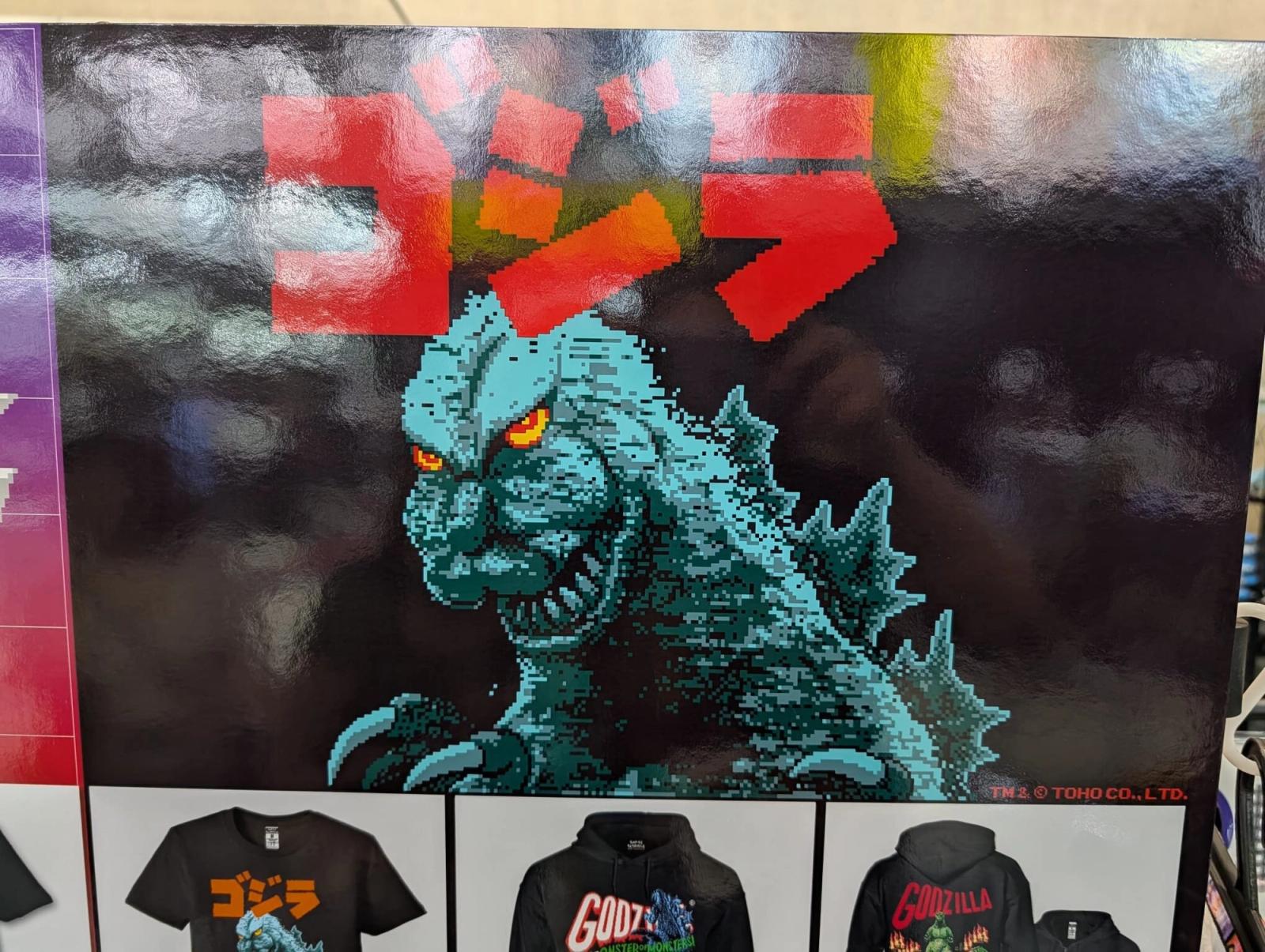 Affisch med Godzilla i pixelgrafik och japansk titeltext ovanför t-shirts och hoodies.