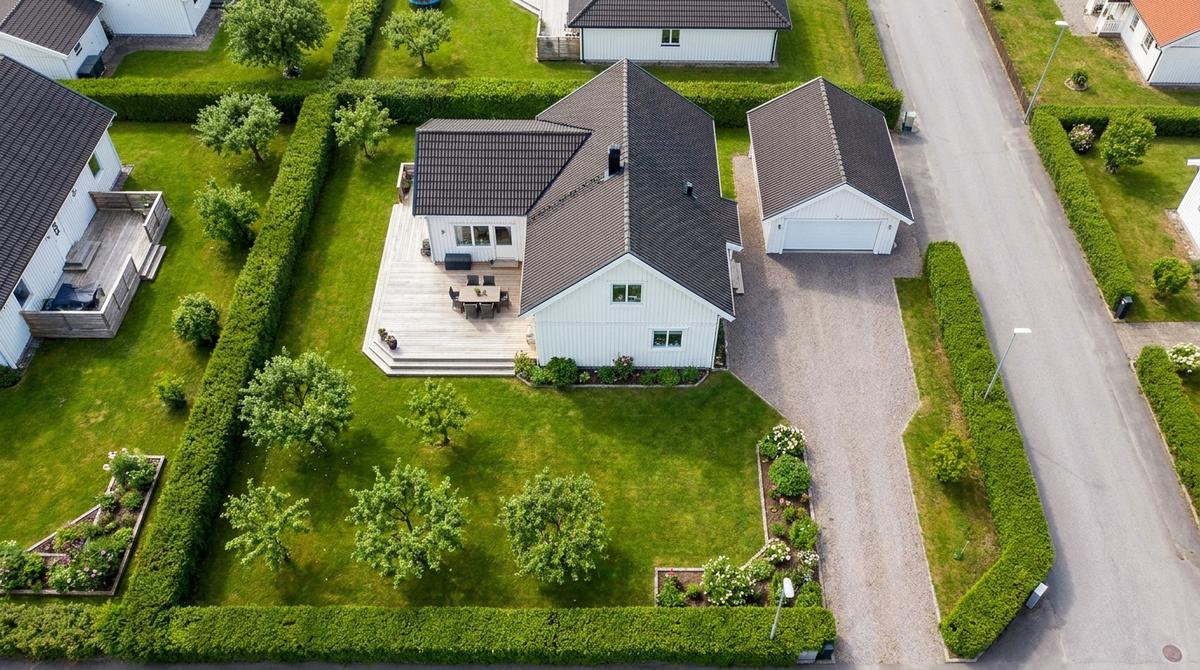 Flygbild högt ovanifrån av en villatomt: vitt hus med mörkt tak, trädäck med utemöbler, garage och grusad infart, gräsmatta med frukträd och häckar som visar tomtens gränser