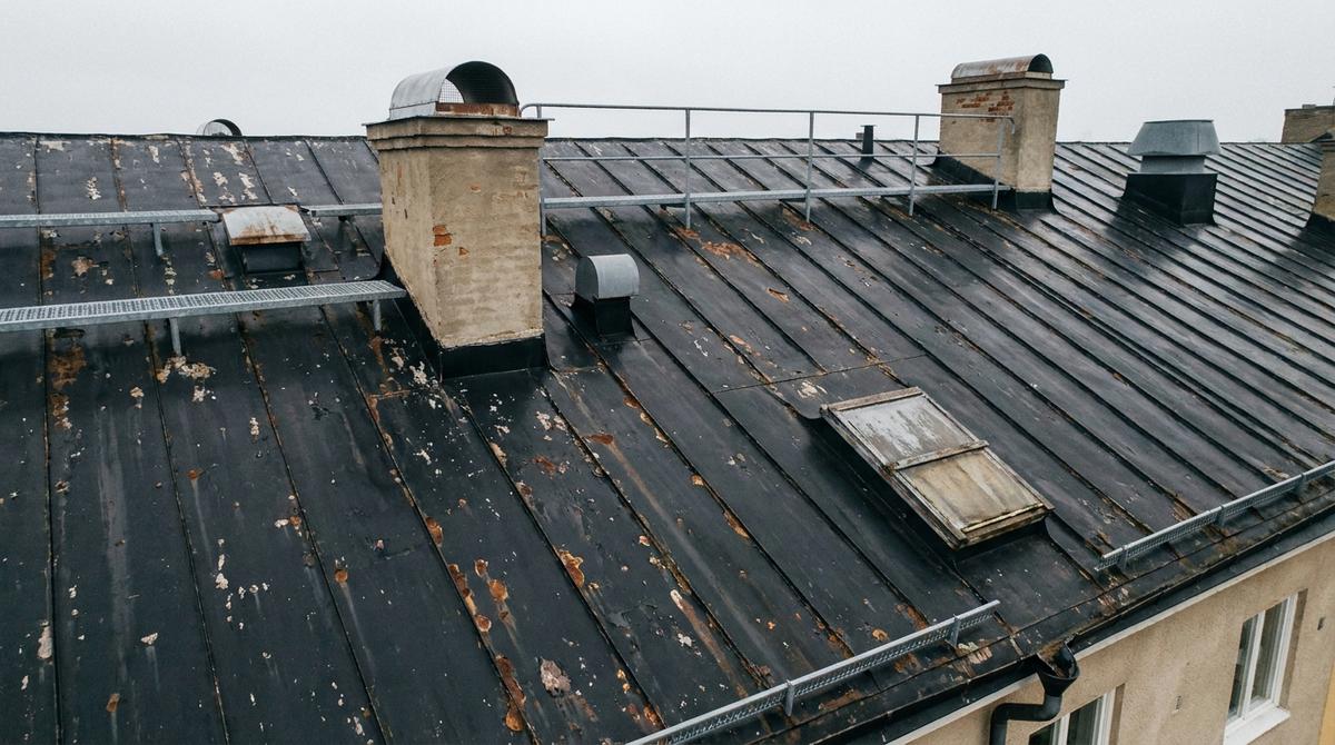 Drönarbild på nära håll av ett svart falsat plåttak med färgsläpp och rostfläckar, två putsade skorstenar med plåthuvar, ventilationshuvar, taklucka, takbrygga och snörasskydd längs takfoten
