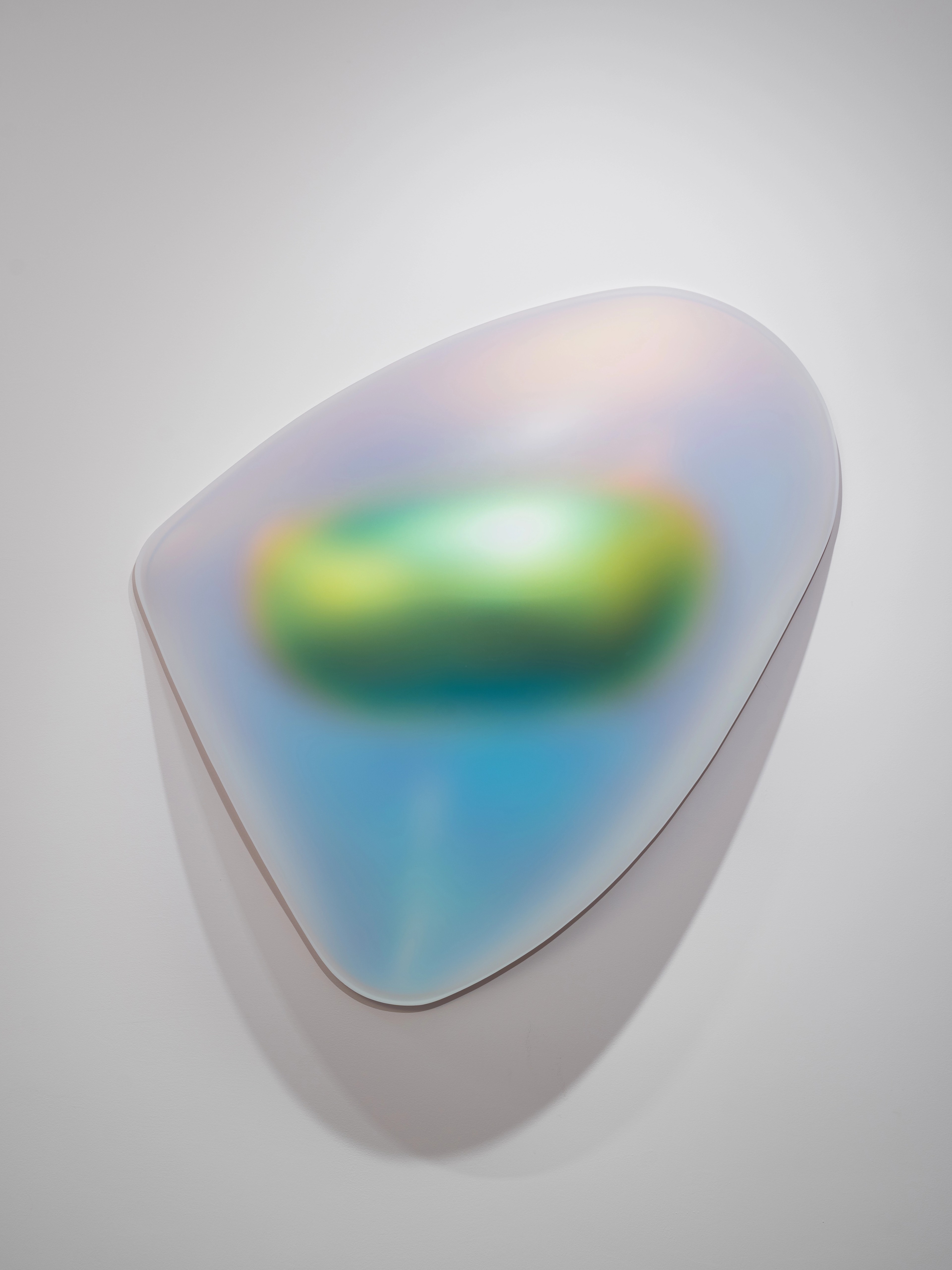 An irregularly shaped wall sculpture with a smooth, semi-translucent surface. The form glows in gradient hues of blue, green, pink, and gold, creating a fluid, futuristic impression reminiscent of a light-diffusing organic object.