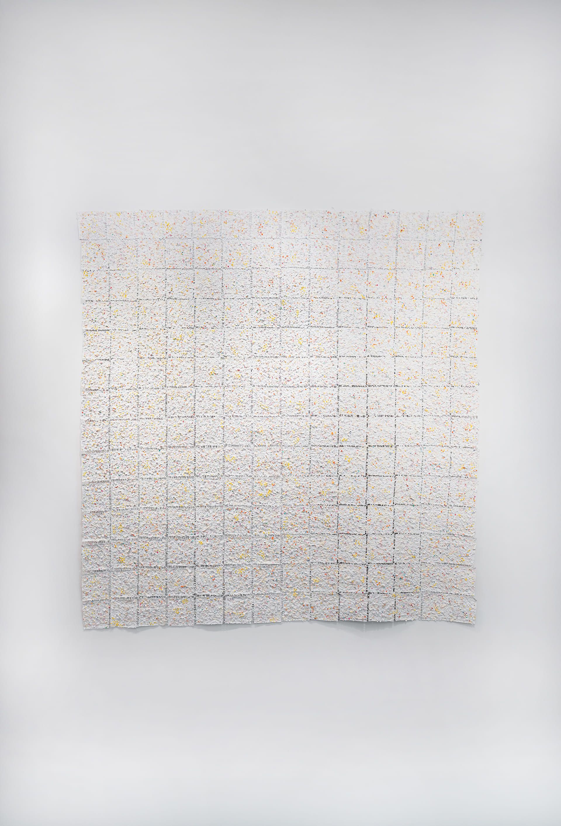A large, textured wall installation composed of a grid of square panels, each covered in a white surface speckled with small dots of color, including yellow, orange, red, and blue. The squares are arranged in neat rows and columns, creating a sense of structure within the organic, handmade texture. The artwork hangs on a white wall under track lighting, with a gray terrazzo baseboard and dark herringbone wood flooring below. The overall effect is delicate, rhythmic, and tactile, inviting close inspection of the detailed, irregular surface.