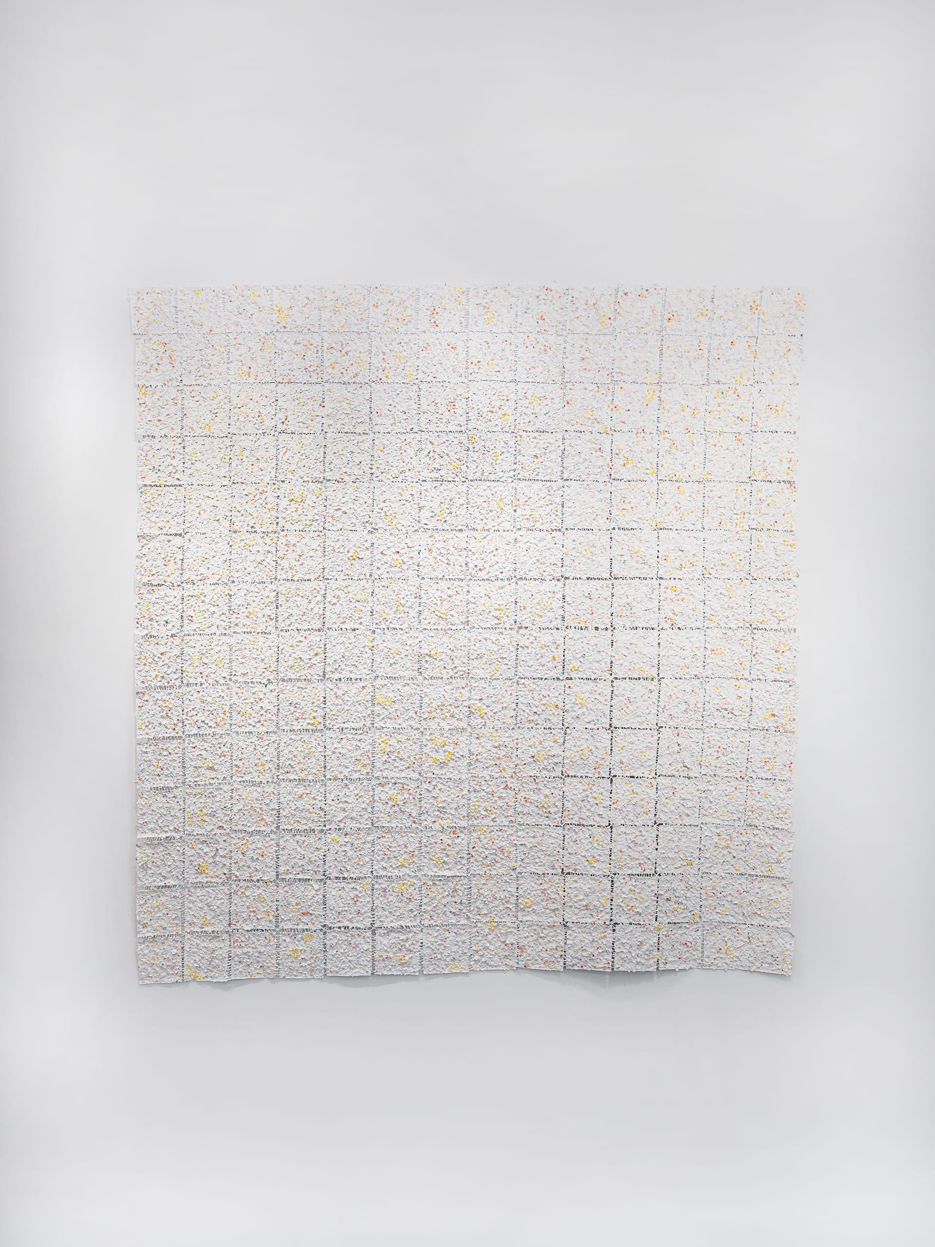 A large, textured wall installation composed of a grid of square panels, each covered in a white surface speckled with small dots of color, including yellow, orange, red, and blue. The squares are arranged in neat rows and columns, creating a sense of structure within the organic, handmade texture. The artwork hangs on a white wall under track lighting, with a gray terrazzo baseboard and dark herringbone wood flooring below. The overall effect is delicate, rhythmic, and tactile, inviting close inspection of the detailed, irregular surface.