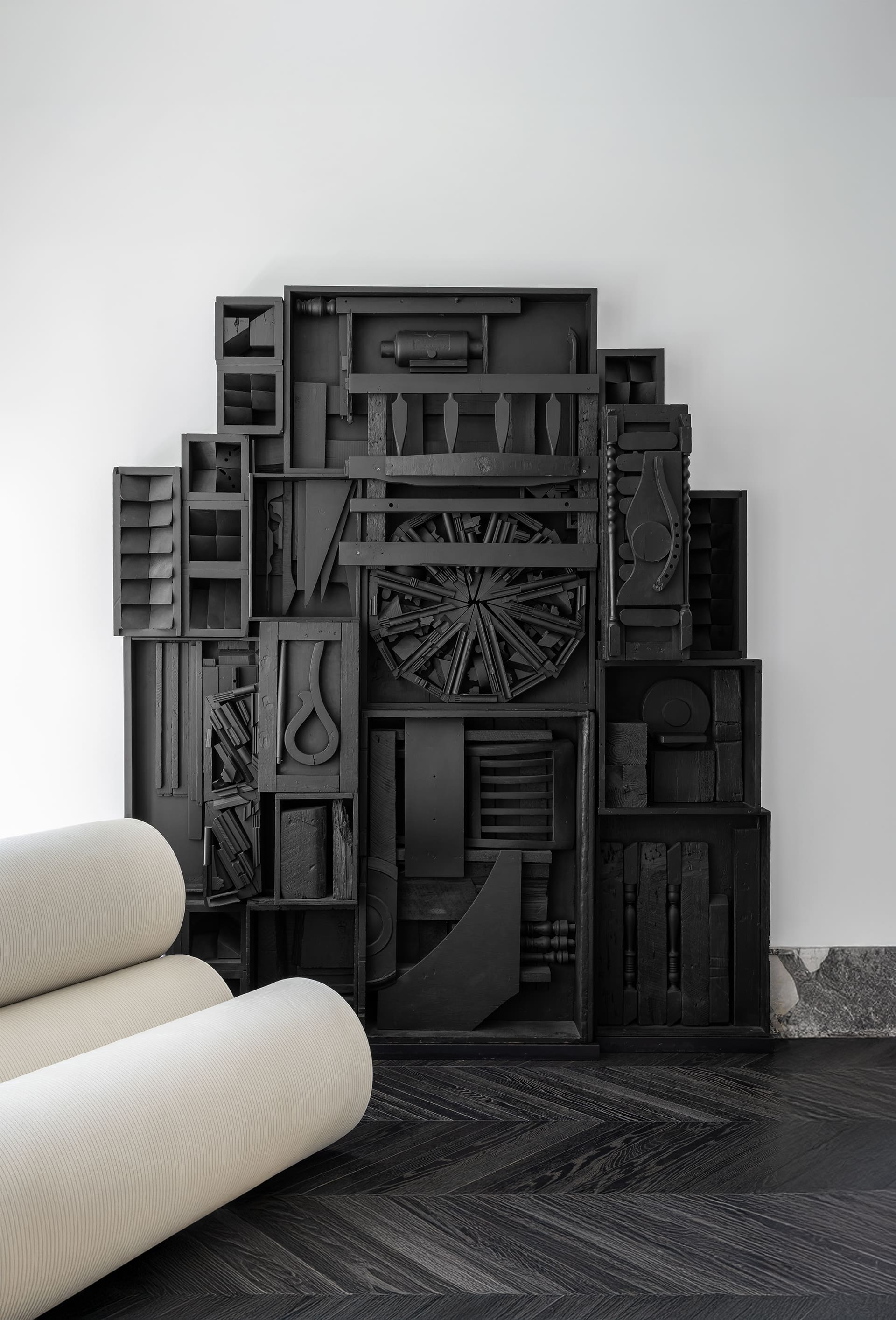 A dramatic black sculptural installation dominates the room, composed of layered geometric forms and intricate textures that create depth and shadow, resembling an abstract architectural relief. Its dark, matte finish contrasts boldly with the light furnishings and vibrant chandelier above.