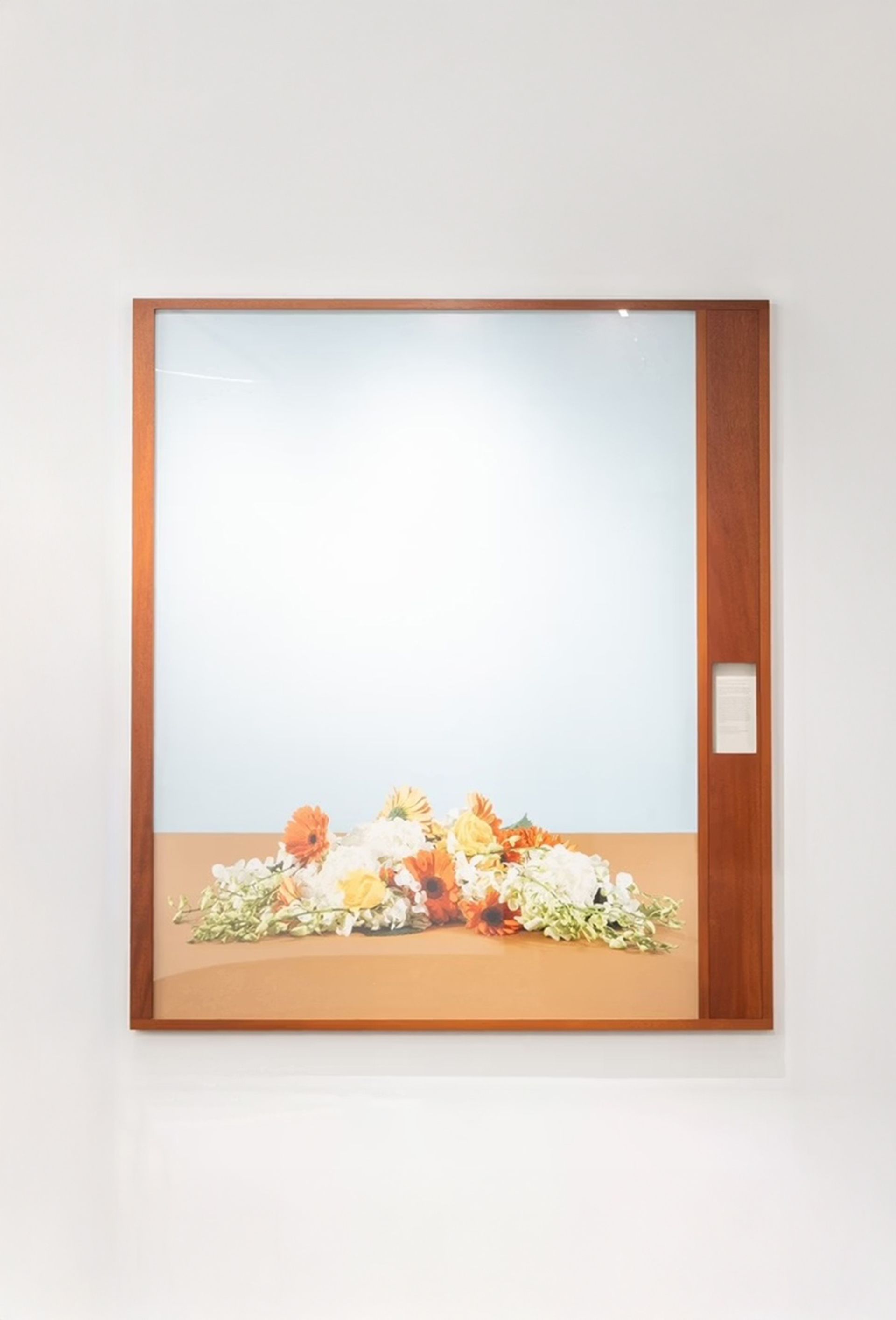 A large framed photograph of a floral arrangement is displayed on a white wall above a stone baseboard. The photograph features a simple, minimalist composition — a cluster of orange, yellow, and white flowers resting on a light brown surface against a pale blue background. The frame is made of warm-toned wood, with a narrow vertical panel on the right side that contains a small descriptive label.