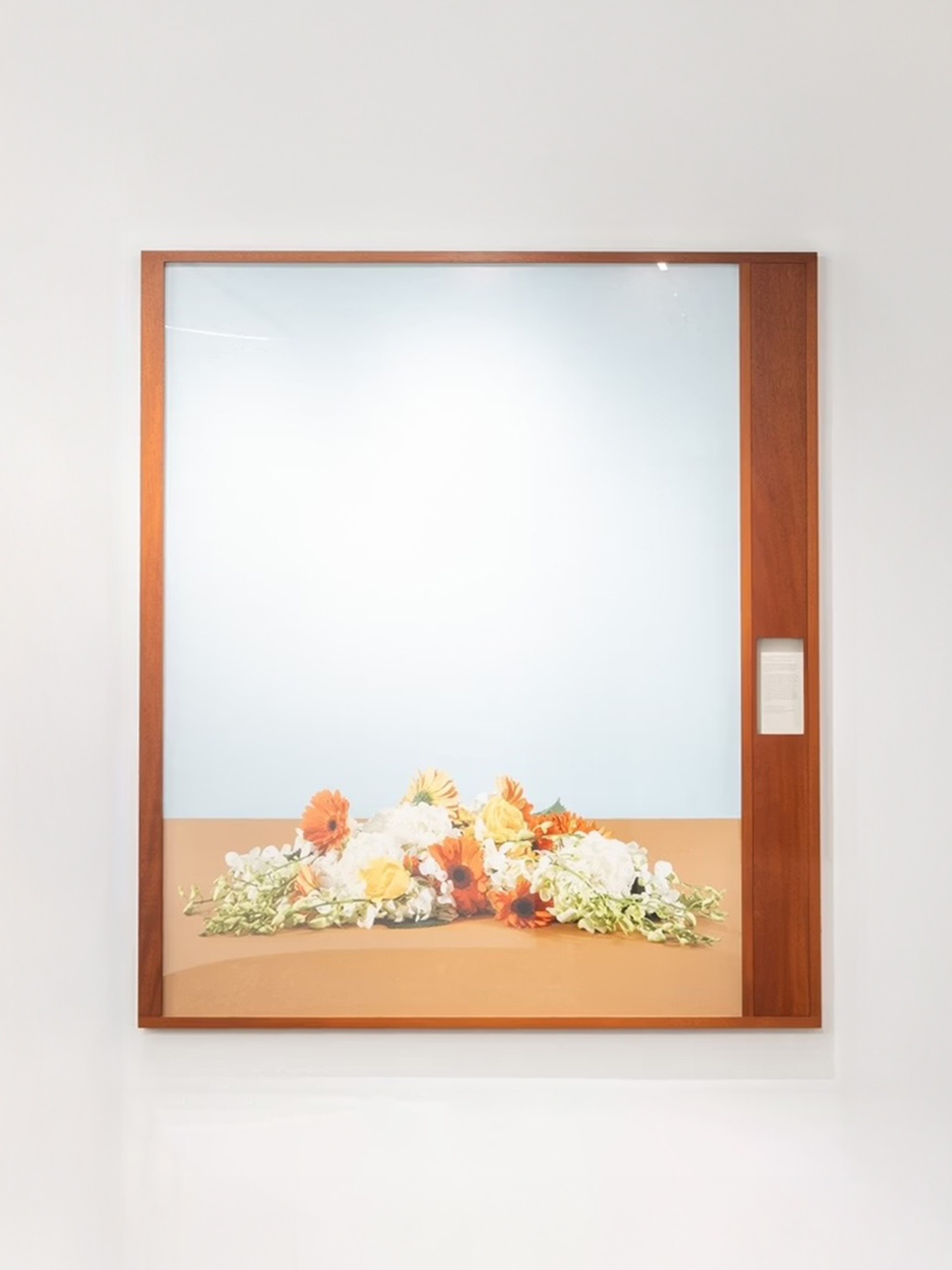 A large framed photograph of a floral arrangement is displayed on a white wall above a stone baseboard. The photograph features a simple, minimalist composition — a cluster of orange, yellow, and white flowers resting on a light brown surface against a pale blue background. The frame is made of warm-toned wood, with a narrow vertical panel on the right side that contains a small descriptive label.