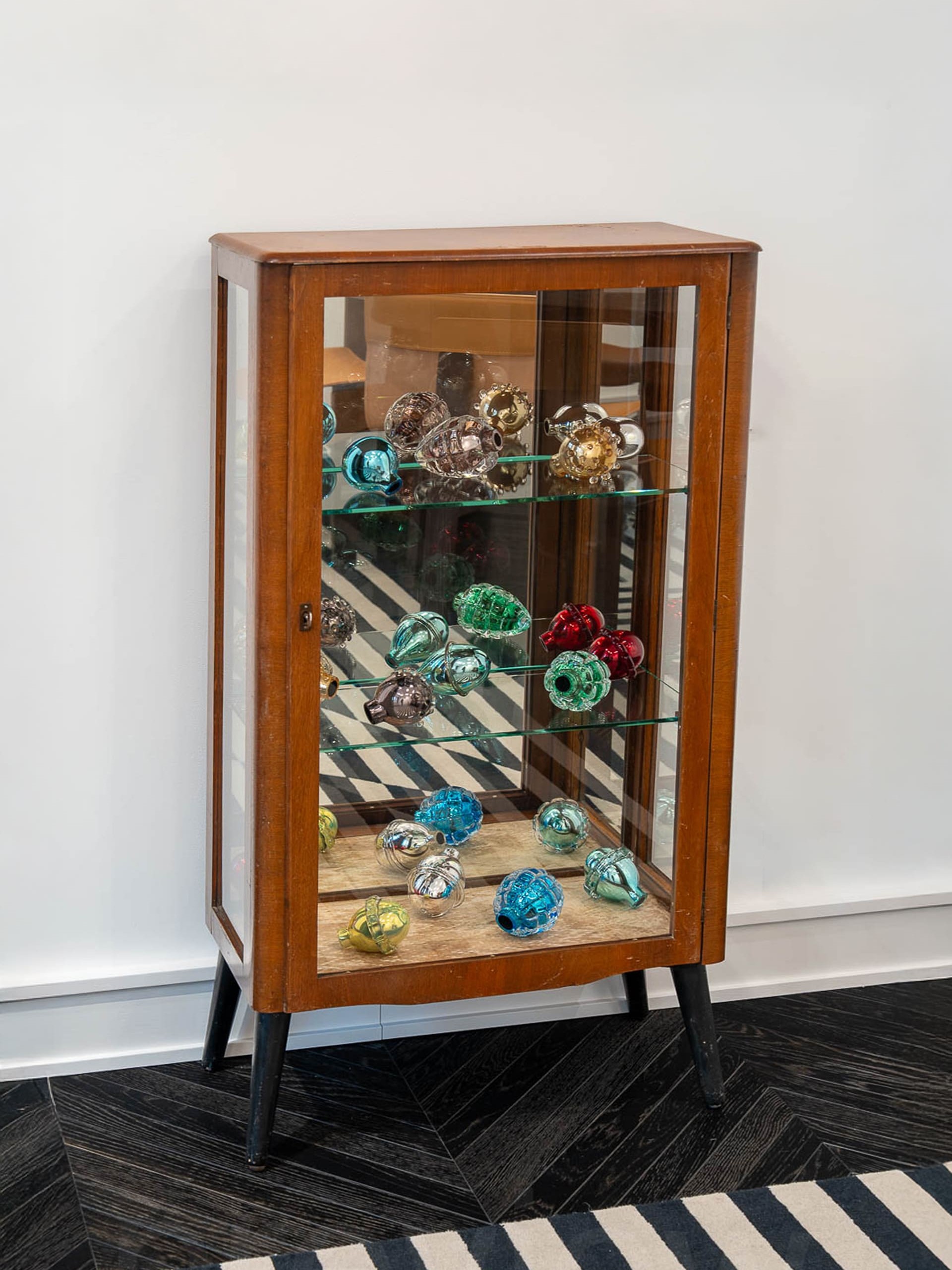 A wooden display cabinet with glass sides and shelves contains an assortment of colorful blown-glass objects. The pieces vary in shape—some rounded, some elongated, some textured with bumps—and come in metallic and translucent colors including silver, gold, red, teal, green, and blue. Each object has a small opening, suggesting they are decorative vessels or ornaments. The glass shelves reflect the vibrant pieces, creating layered reflections within the case.
