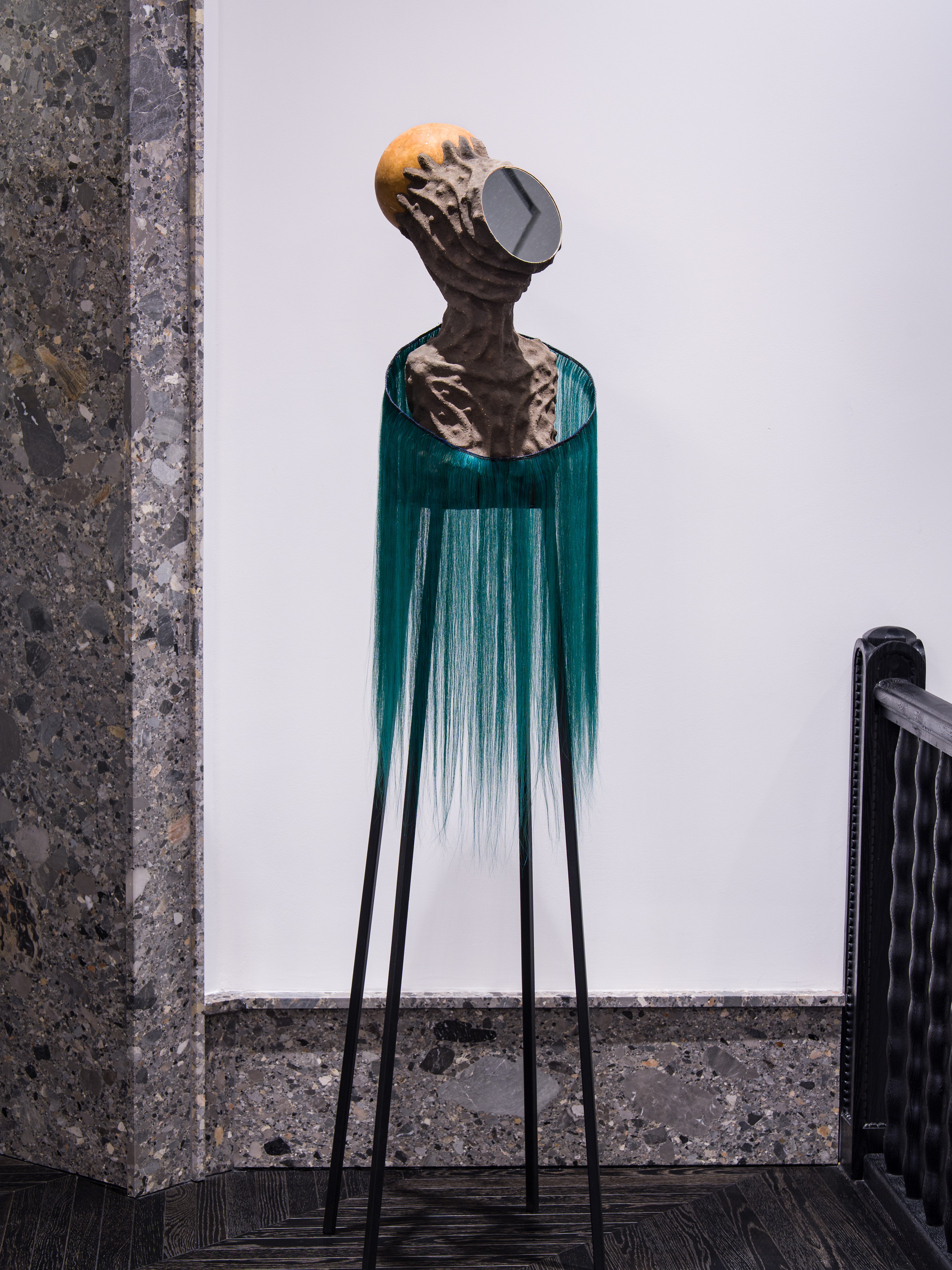 A tall, slender sculpture resembling a human bust with an organic, textured surface and teal fibers cascading downward like hair. The head features a circular reflective disc, giving the piece a futuristic yet ritualistic presence.