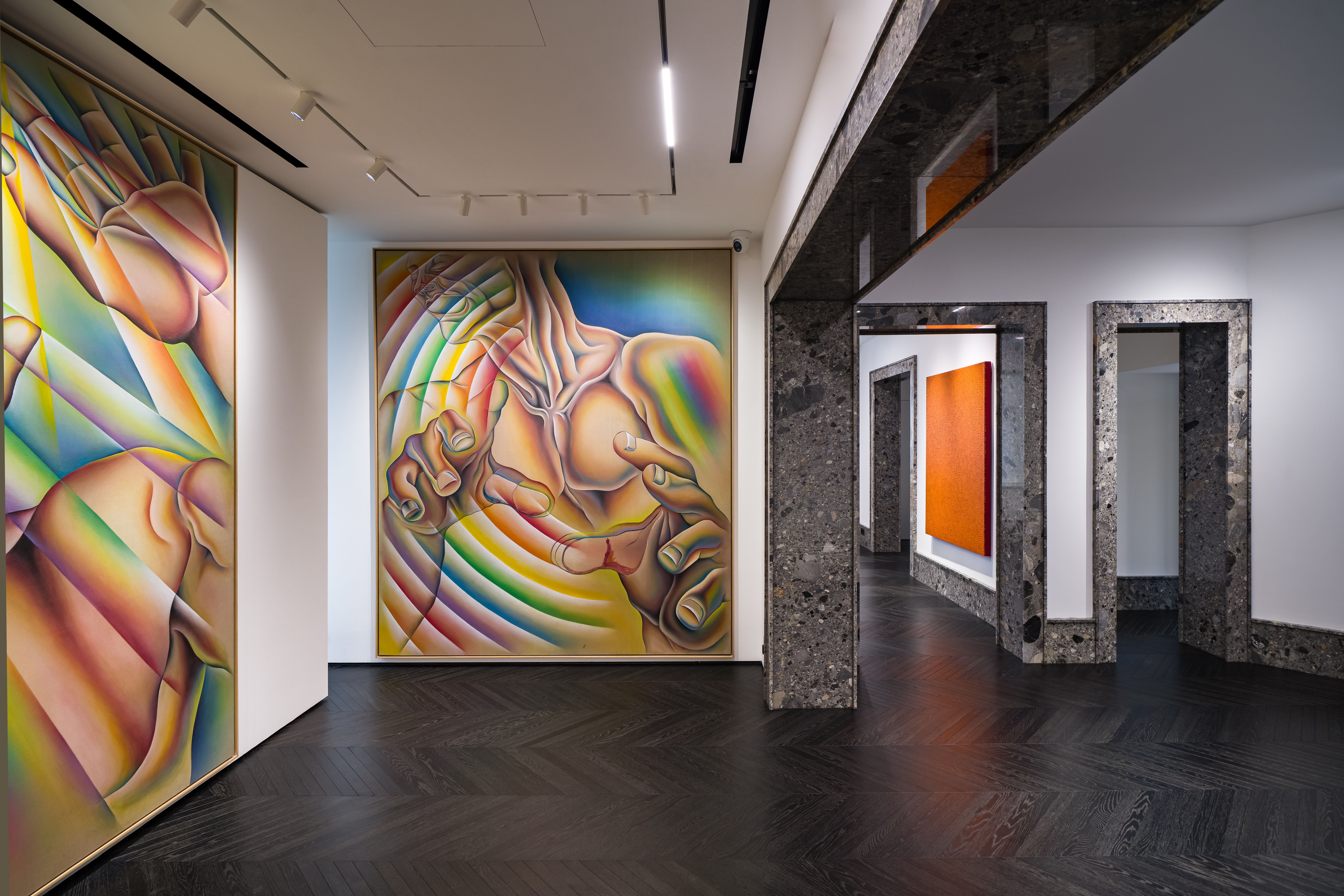 An interior gallery space with large, vividly colored paintings featuring abstracted human forms in rainbow gradients. The room has dark herringbone wood floors, white walls, and polished stone door frames leading to adjoining exhibition rooms with additional artworks.