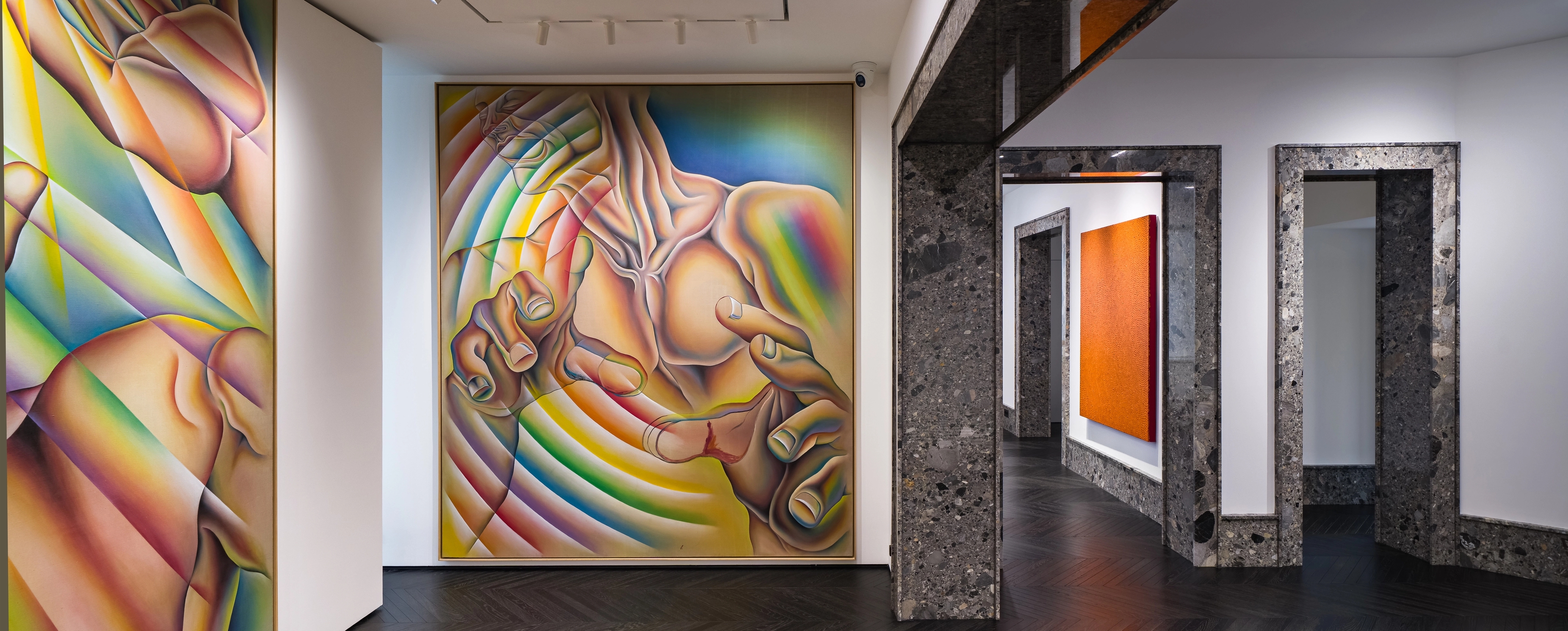 An interior gallery space with large, vividly colored paintings featuring abstracted human forms in rainbow gradients. The room has dark herringbone wood floors, white walls, and polished stone door frames leading to adjoining exhibition rooms with additional artworks.