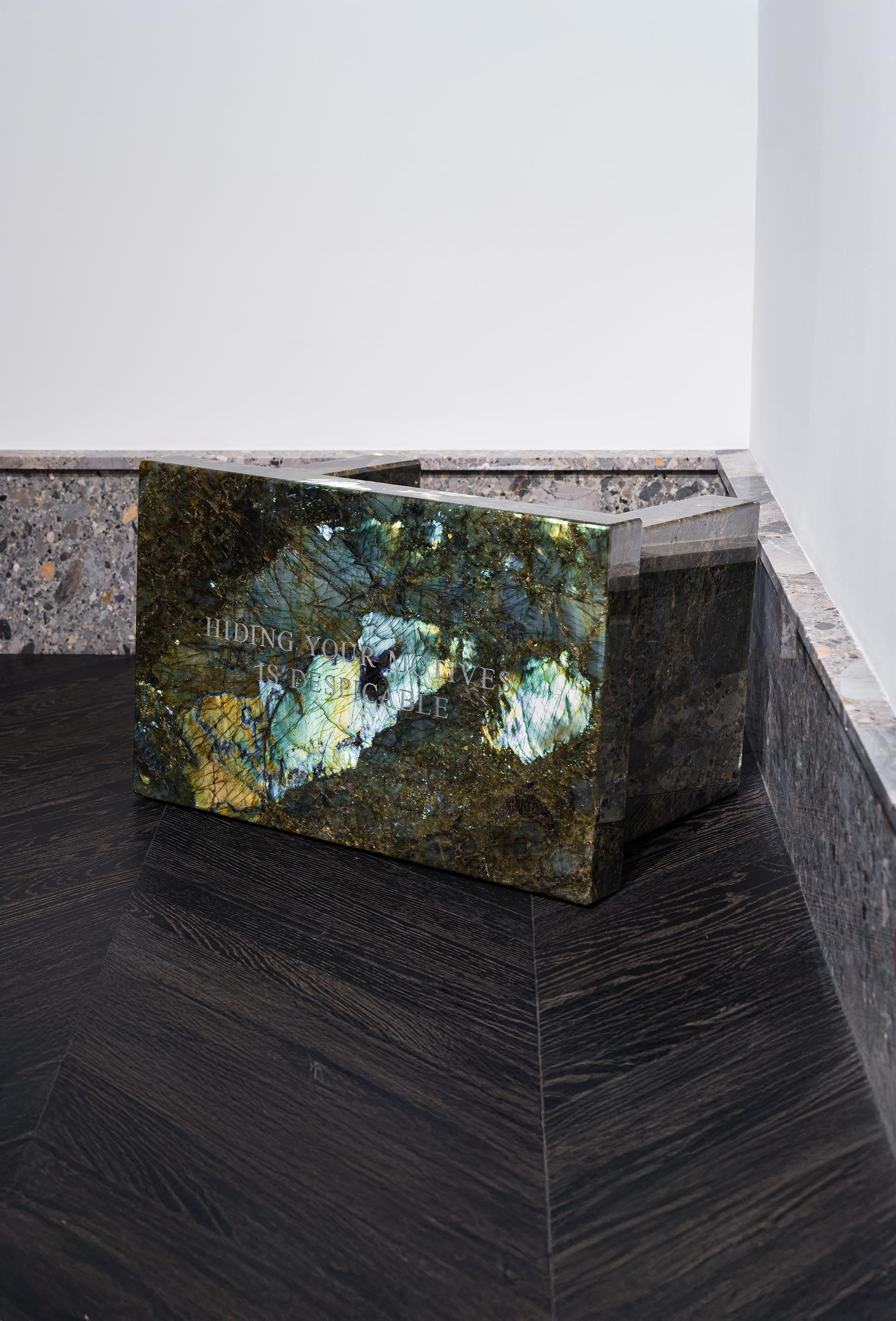 A rectangular sculptural block made of iridescent dark green and blue labradorite stone, positioned in the corner of a gallery space with stone trim and dark wood flooring. The polished surface reflects light, revealing flashes of gold and turquoise. Engraved text on the front reads, “HIDING YOUR MOTIVES IS DESPICABLE.”