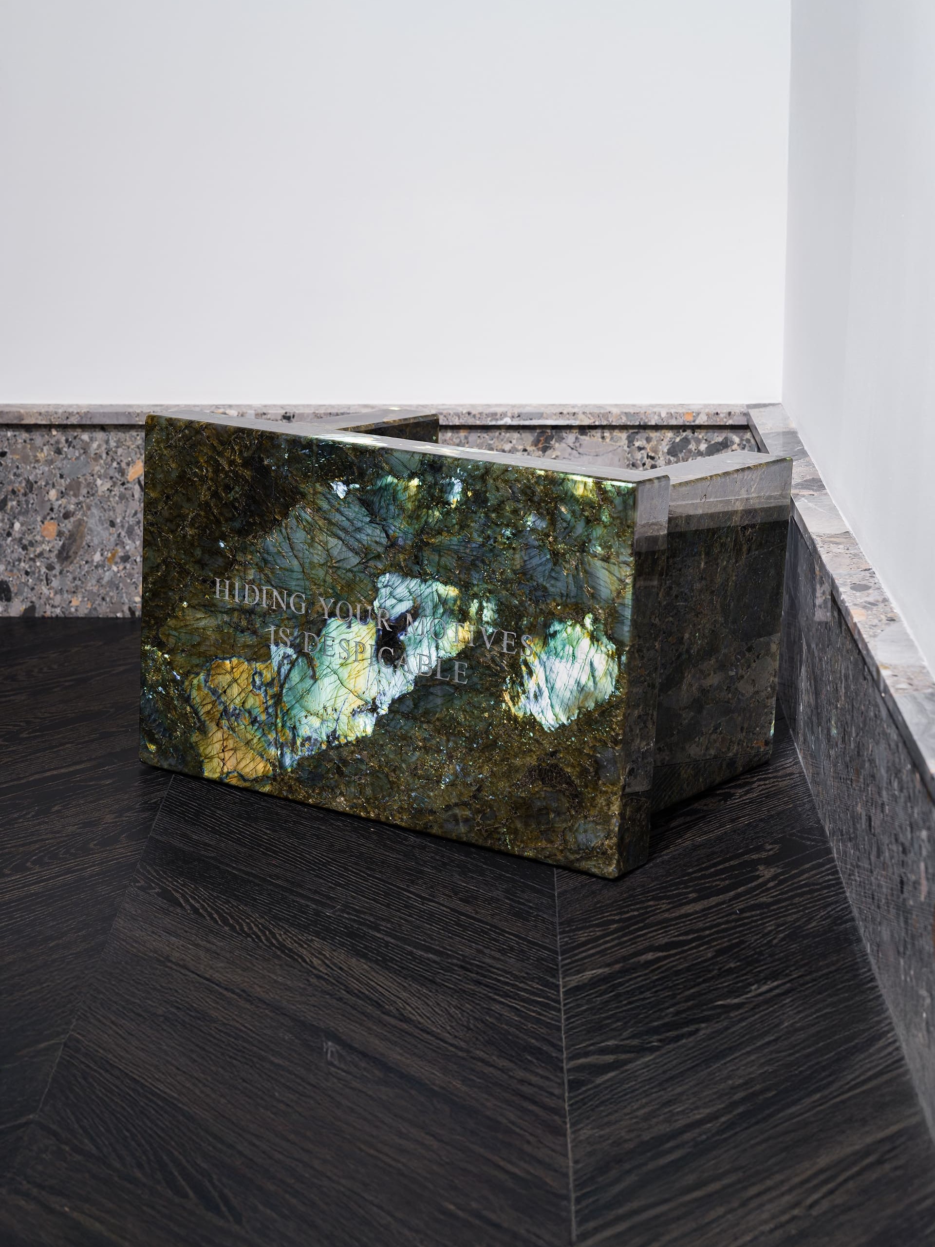 A rectangular sculptural block made of iridescent dark green and blue labradorite stone, positioned in the corner of a gallery space with stone trim and dark wood flooring. The polished surface reflects light, revealing flashes of gold and turquoise. Engraved text on the front reads, “HIDING YOUR MOTIVES IS DESPICABLE.”