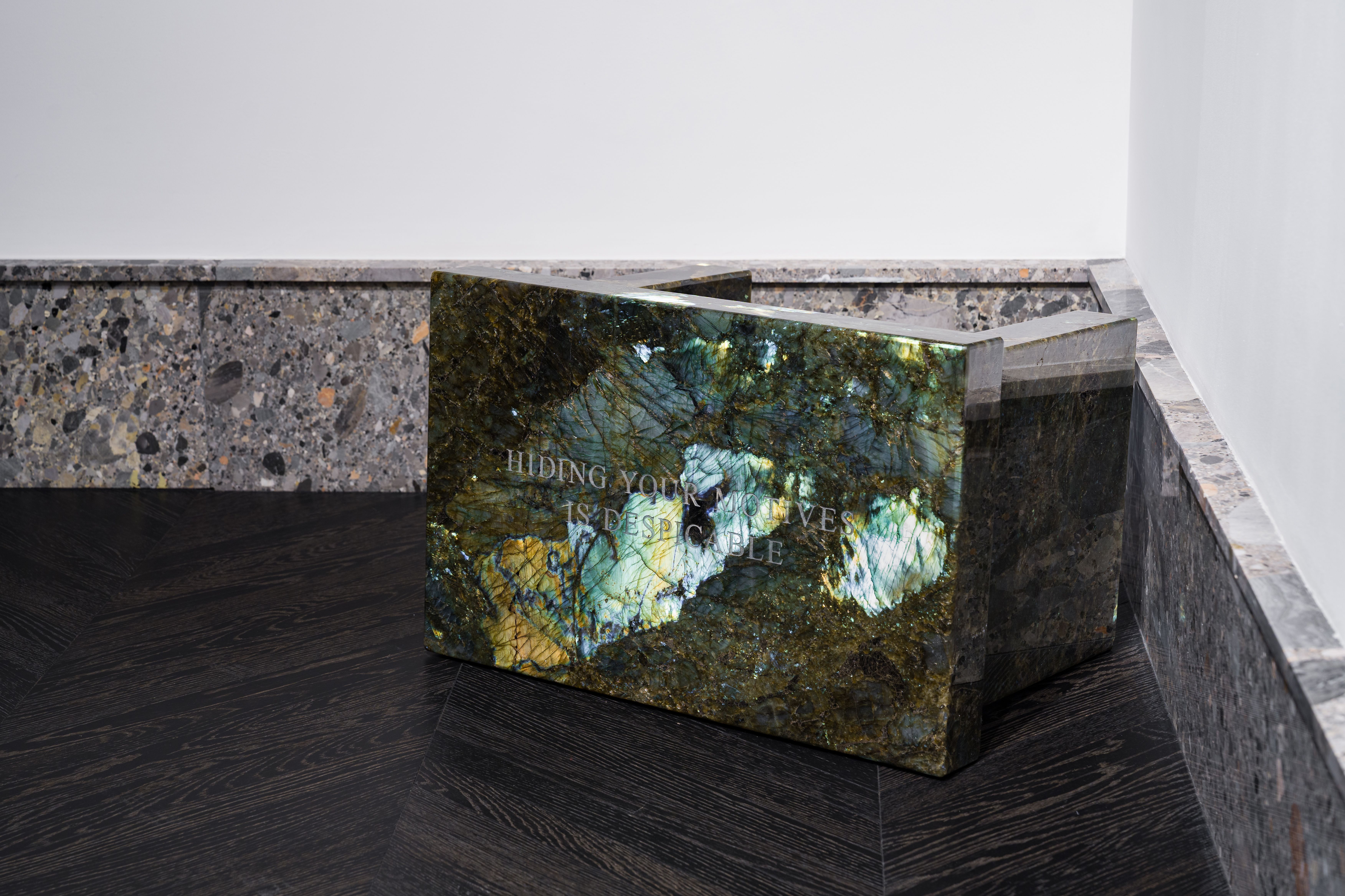 Close-up of a polished iridescent green stone sculpture inscribed with the words “HIDING YOUR MOTIVES IS DESPICABLE.” The piece rests on a dark wood floor beside a wall lined with gray stone, reflecting light that highlights its natural mineral textures.