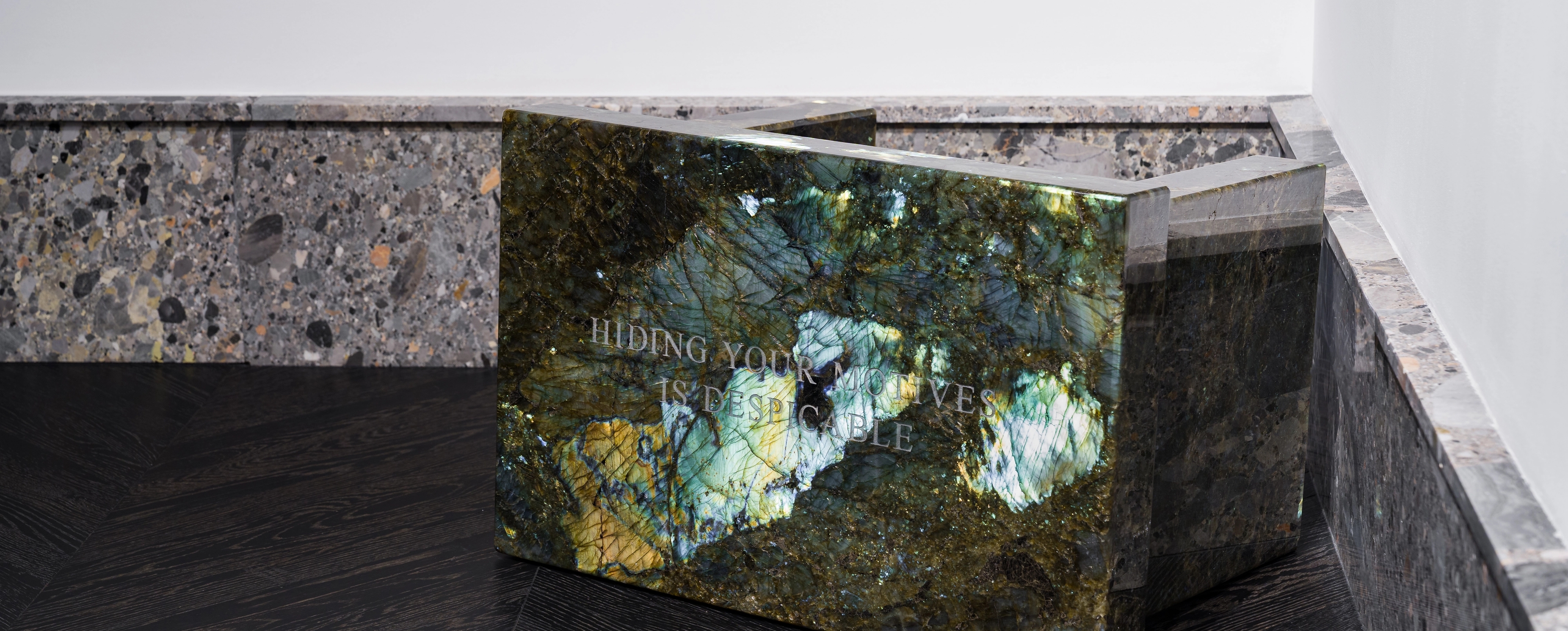 Close-up of a polished iridescent green stone sculpture inscribed with the words “HIDING YOUR MOTIVES IS DESPICABLE.” The piece rests on a dark wood floor beside a wall lined with gray stone, reflecting light that highlights its natural mineral textures.
