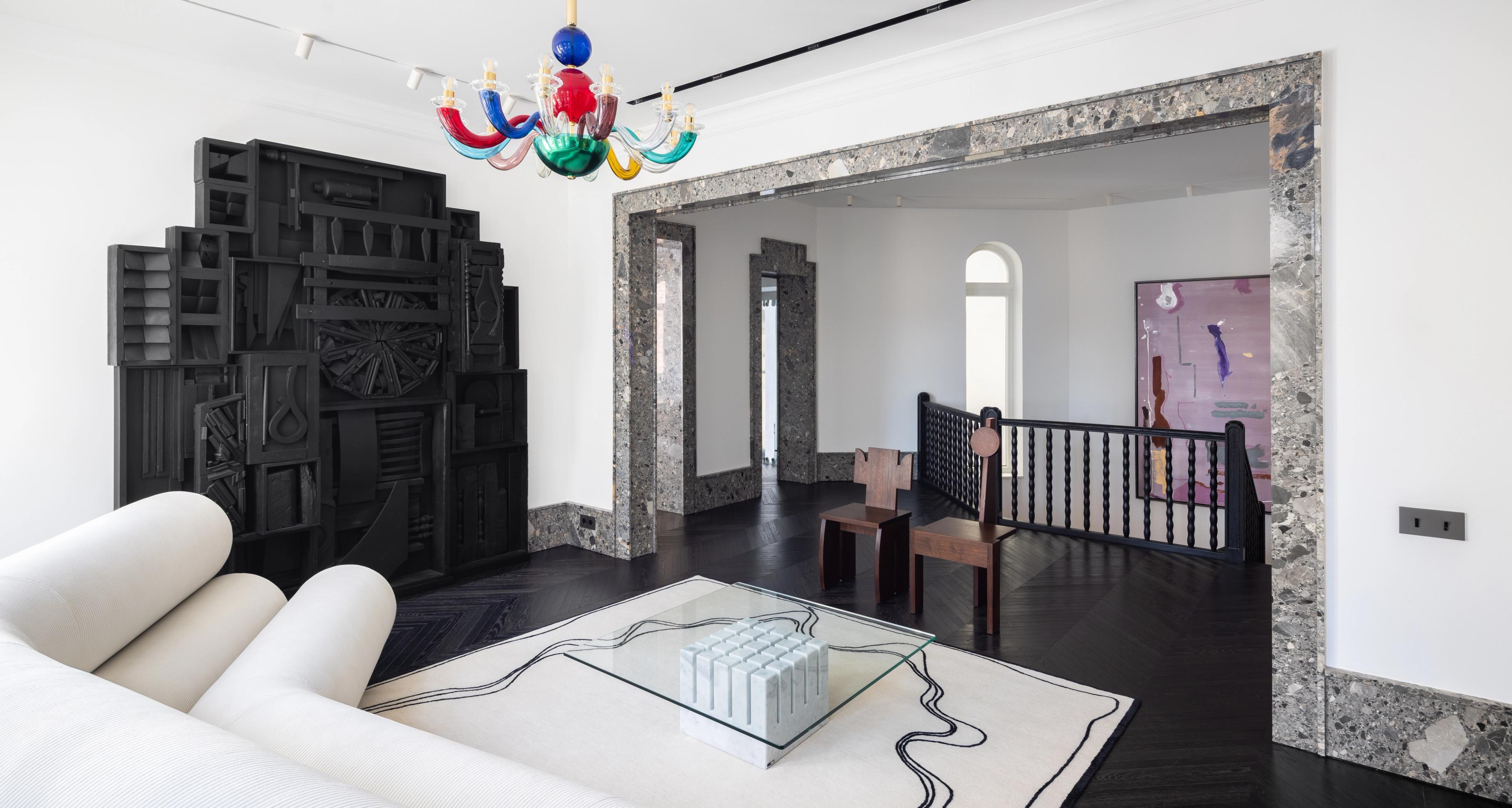A modern lounge area within the gallery, featuring a sculptural white sofa, glass coffee table with a marble base, and black geometric wall installation. The room is framed with stone doorways and accented by a colorful chandelier and contemporary artworks.