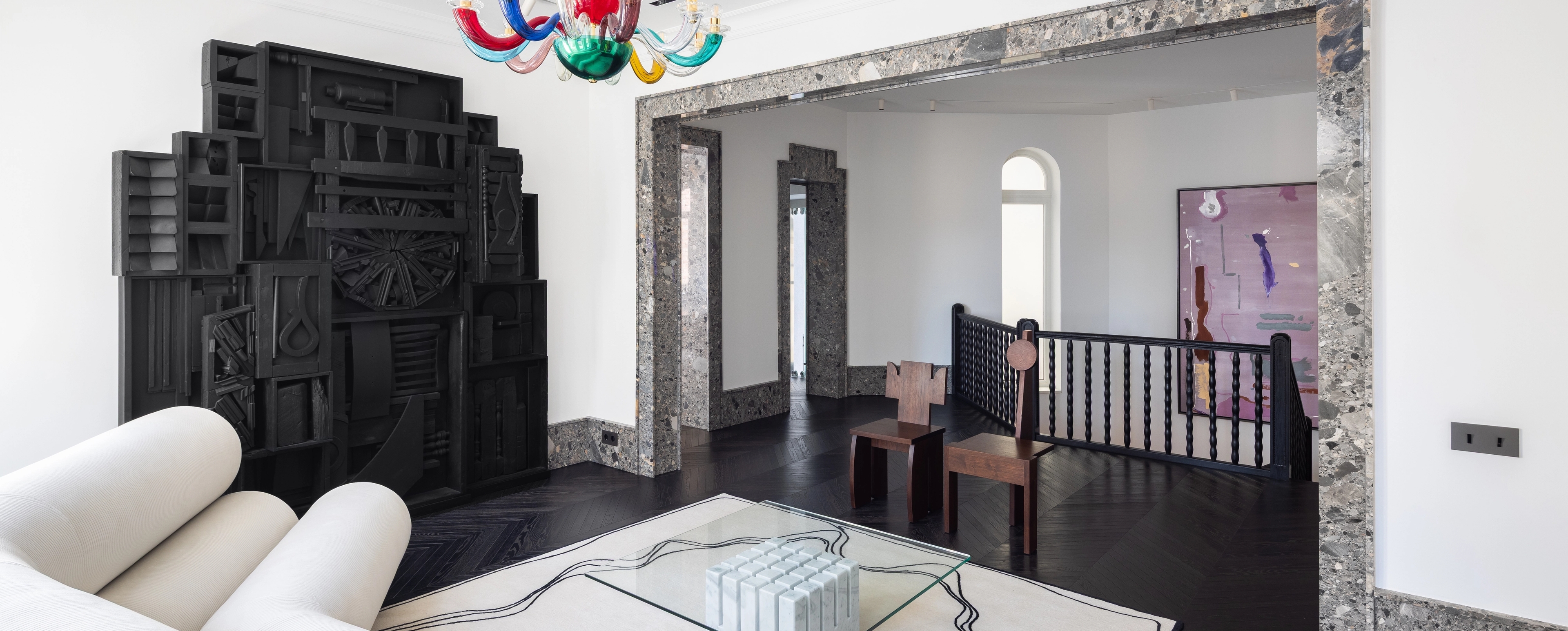 A modern lounge area within the gallery, featuring a sculptural white sofa, glass coffee table with a marble base, and black geometric wall installation. The room is framed with stone doorways and accented by a colorful chandelier and contemporary artworks.