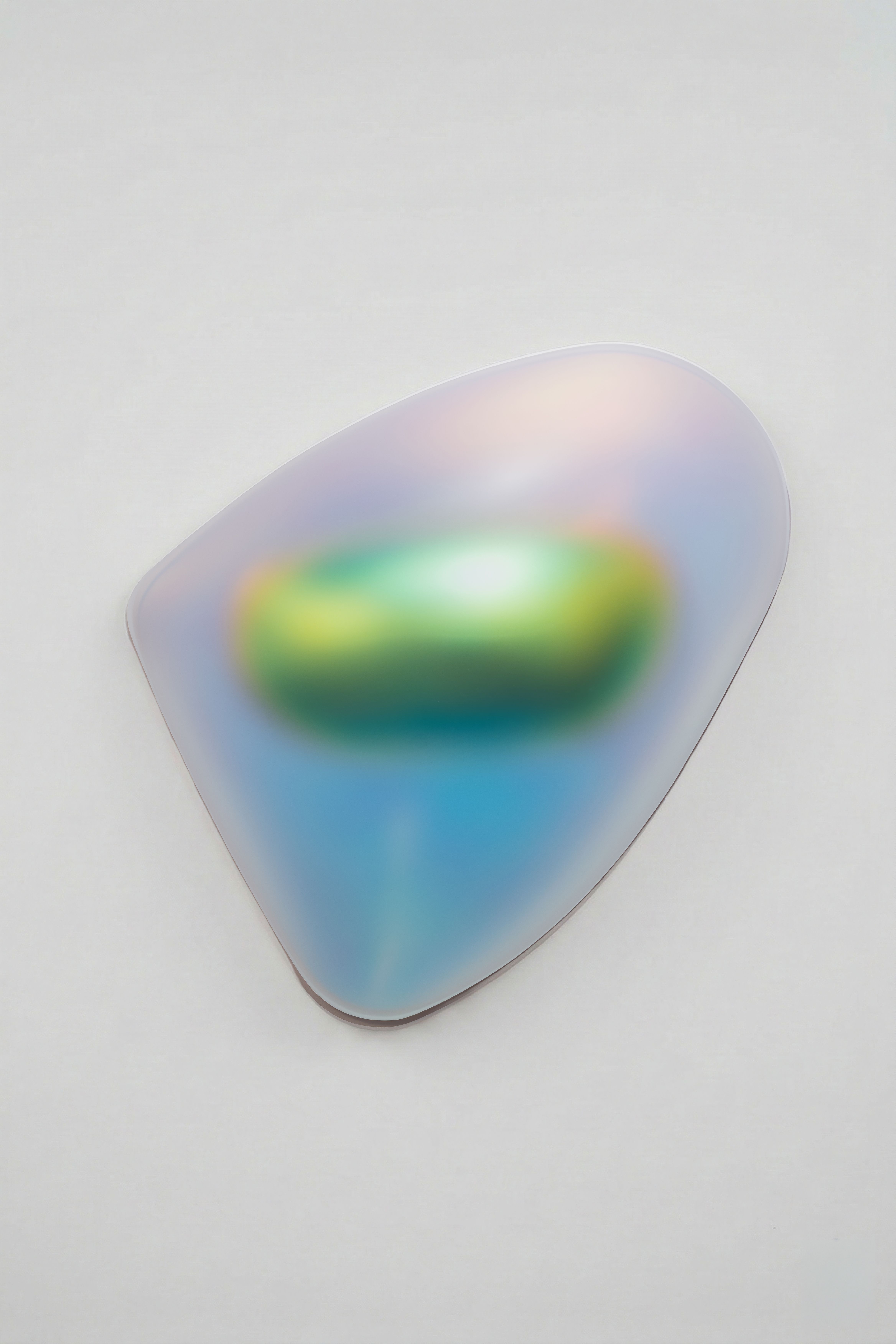 An abstract wall sculpture with a smooth, organic shape resembling a rounded triangle. The surface has an iridescent, translucent finish that shifts in color from green and yellow at the center to blue, violet, and soft pink toward the edges. The artwork appears to glow subtly against a white gallery wall, creating a gentle shadow beneath it.